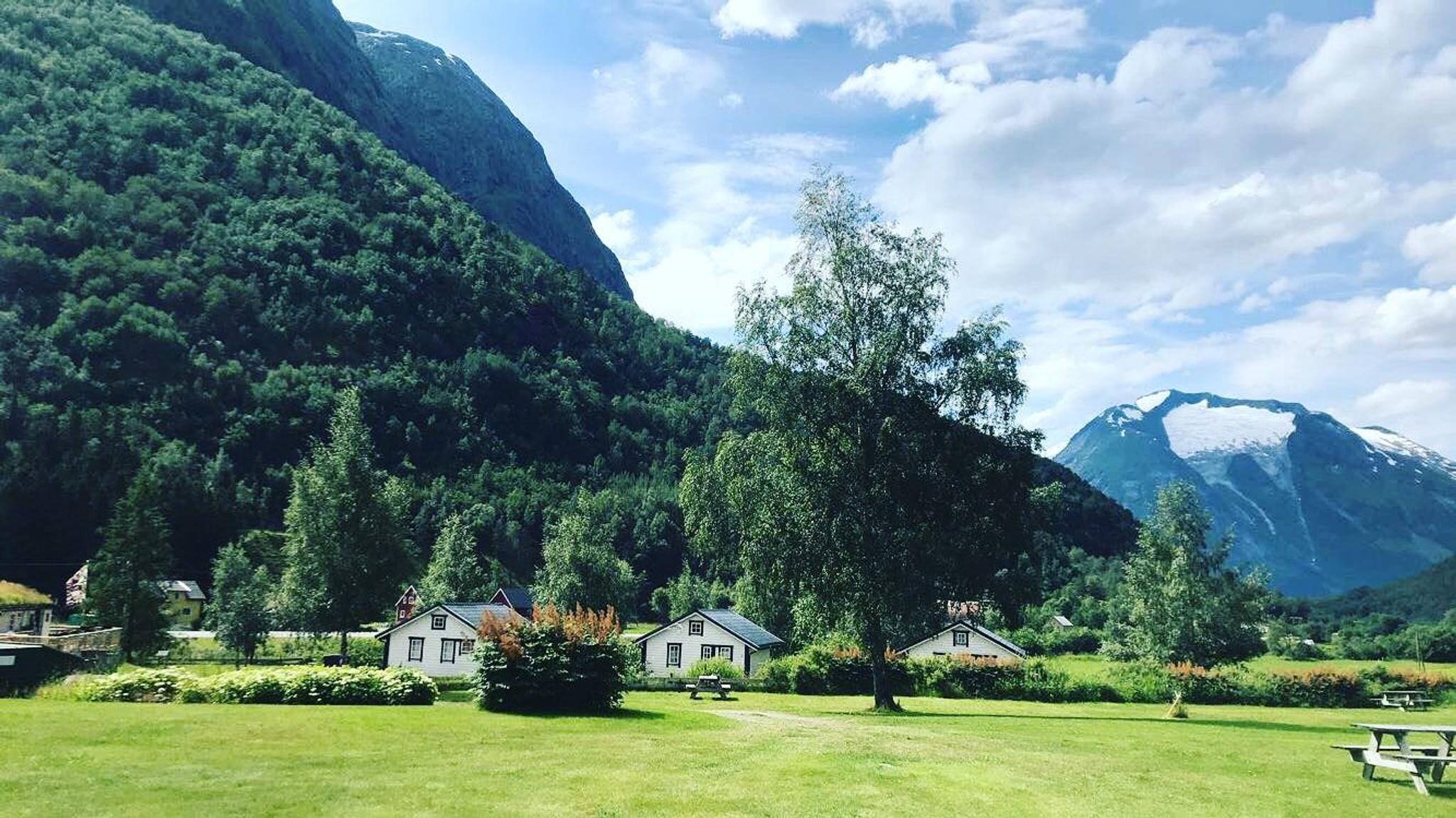 Grøn eng med små kvite hus og tre i forgrunnen, skogkledd fjell og snøtopp i bakgrunnen under skyer..