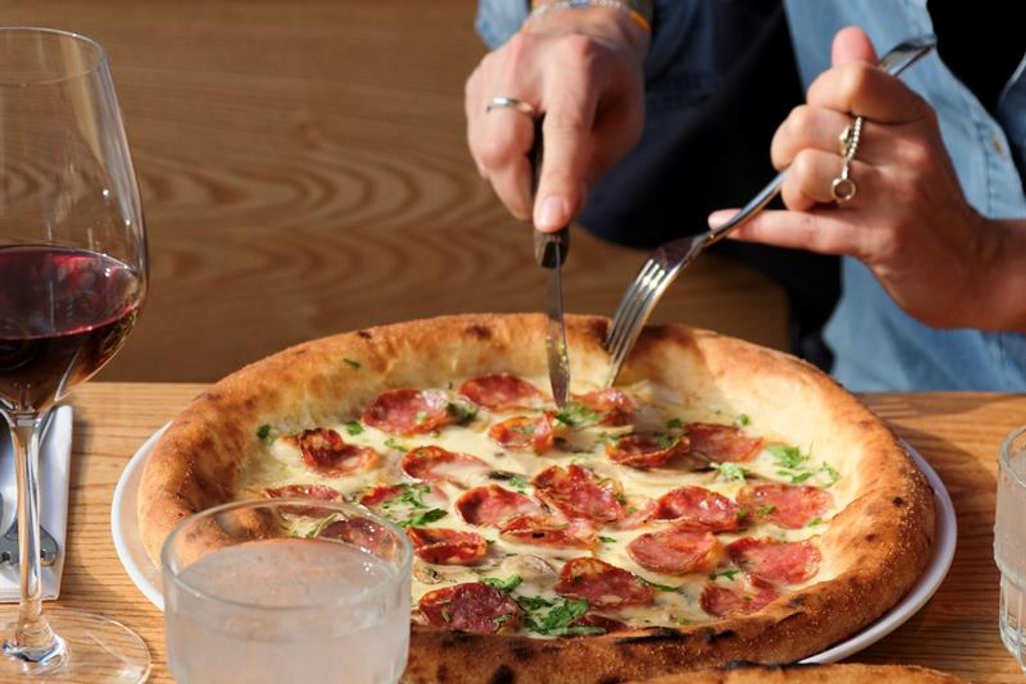 Detail of hands cutting a pizza with a knife and fork, alongside a glass of red wine.