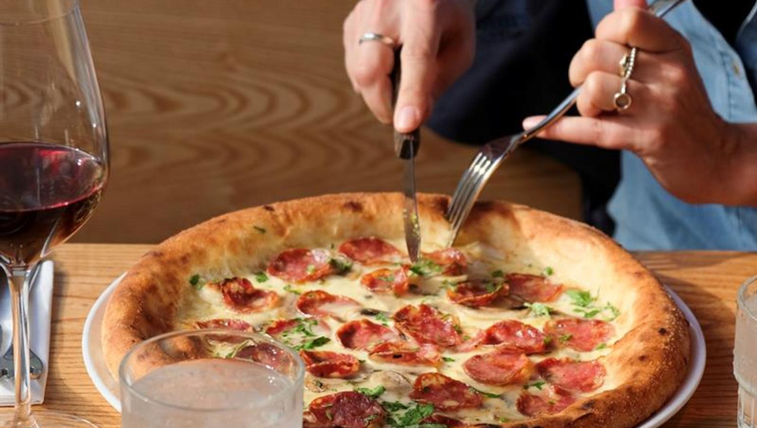 Detail of hands cutting a pizza with a knife and fork, alongside a glass of red wine.