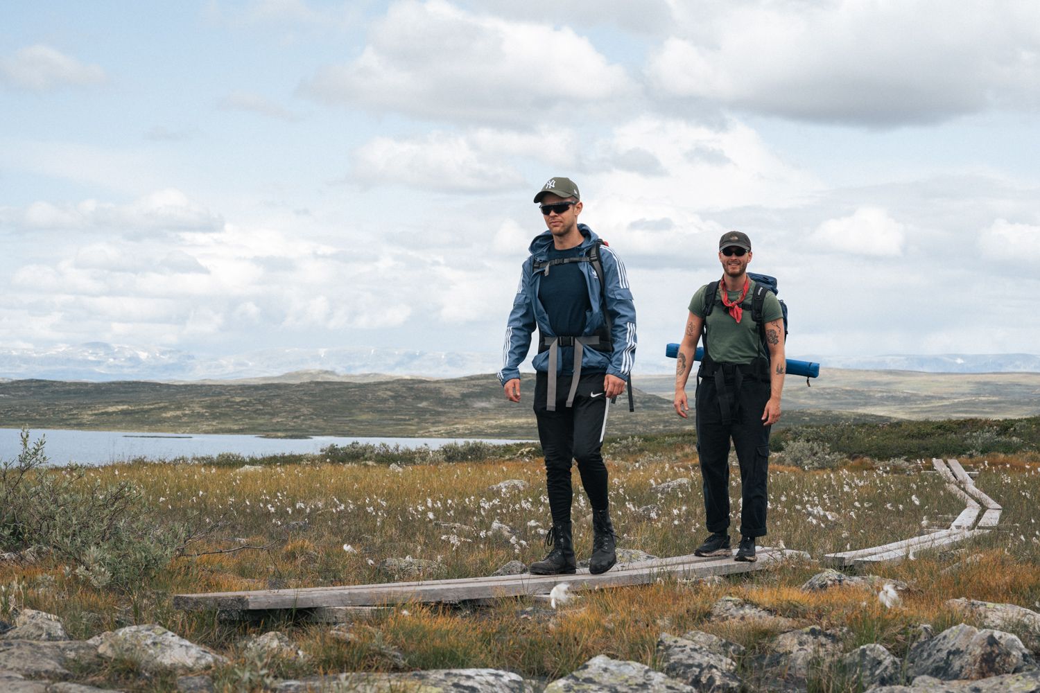 Two hikers are walking on wooden logs therough marsh landscape.