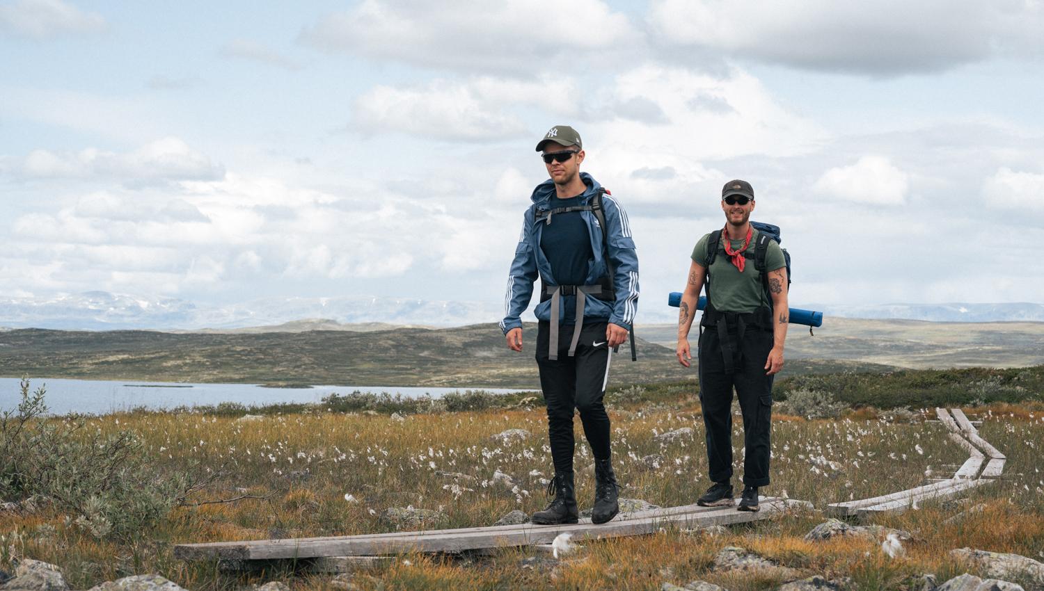 Two hikers are walking on wooden logs therough marsh landscape.