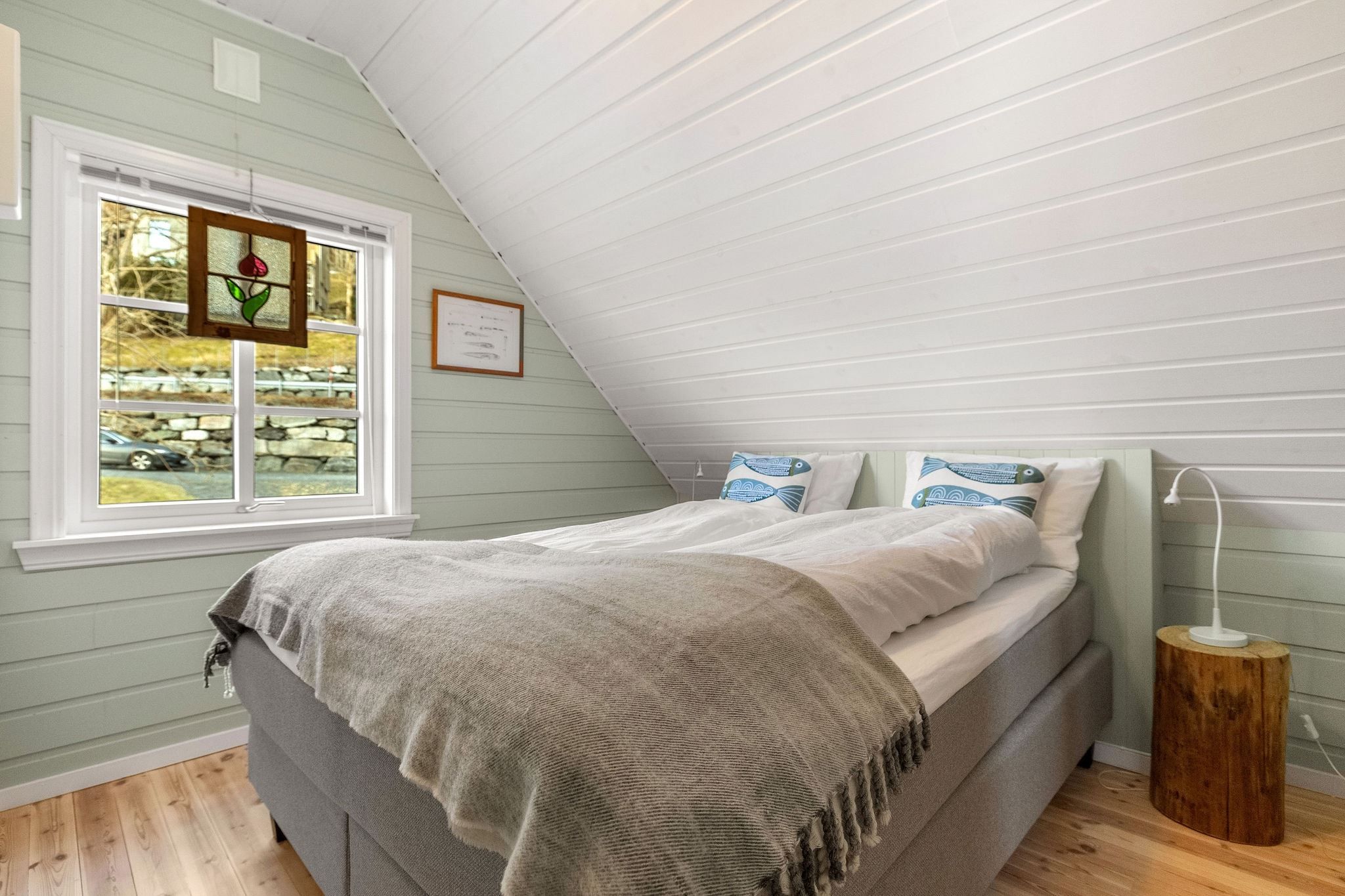 Small attic bedroom with a double bed under a sloped white ceiling, window with stained glass, and a bedside lamp at the Seahouse at Hornelen Panorama in Bremanger...