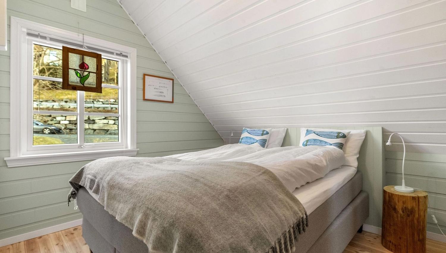 Small attic bedroom with a double bed under a sloped white ceiling, window with stained glass, and a bedside lamp at the Seahouse at Hornelen Panorama in Bremanger...