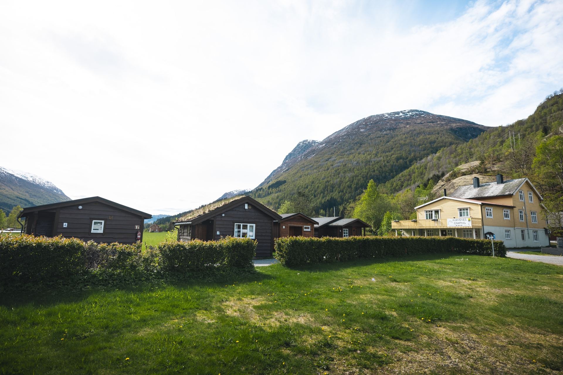 Wooden cabins and a light-colored lodge sit behind hedges on a grassy lawn, with forested mountains under a bright sky..