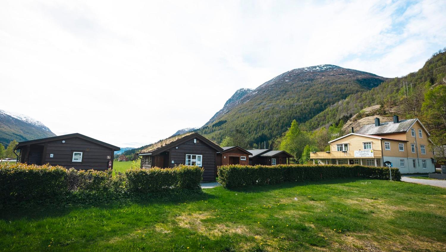 Wooden cabins and a light-colored lodge sit behind hedges on a grassy lawn, with forested mountains under a bright sky..