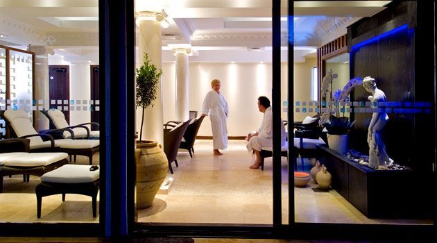 Interior view of a spa lounge seen through glass doors, with two people in robes near chairs and a water feature.