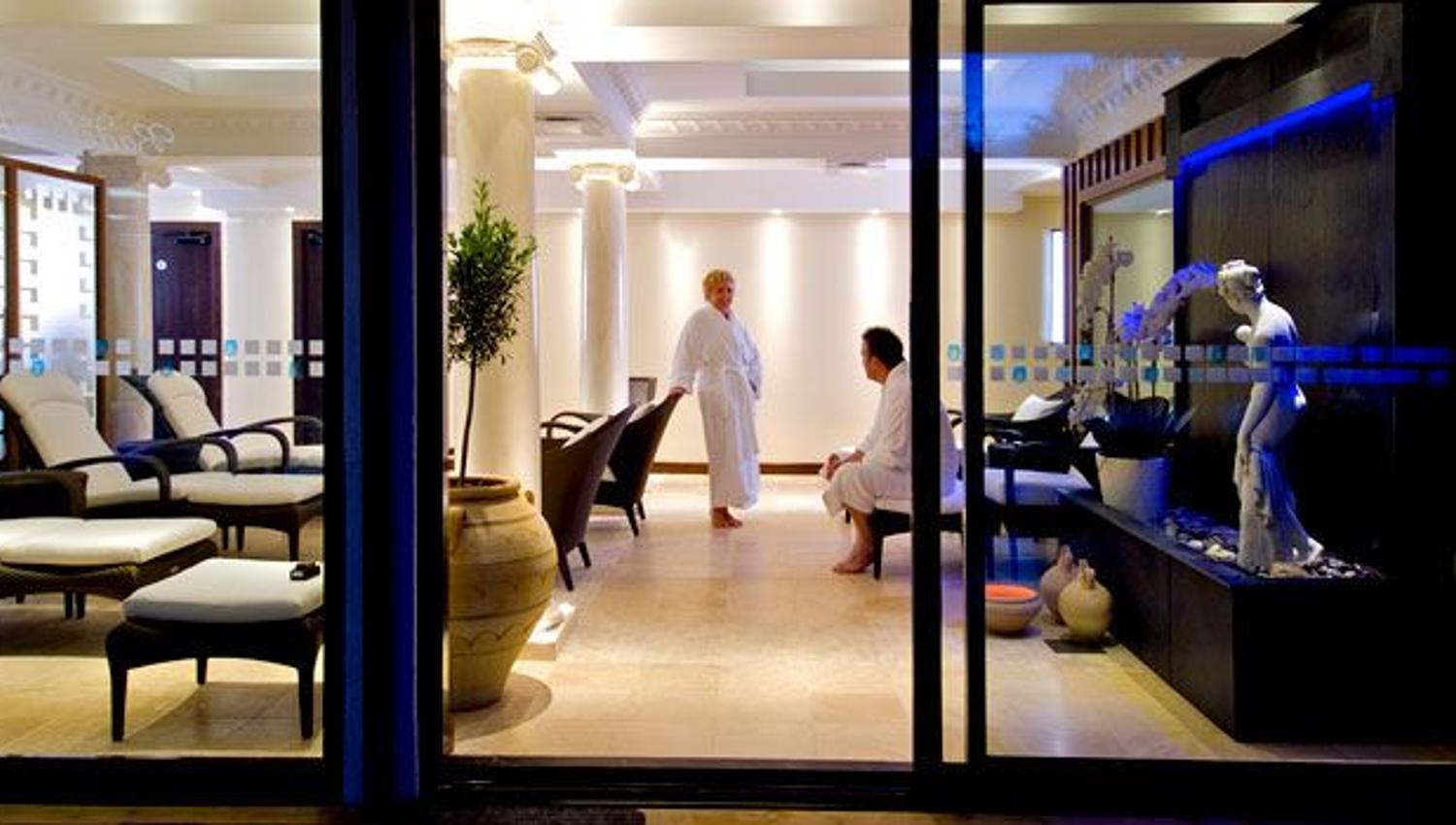 Interior view of a spa lounge seen through glass doors, with two people in robes near chairs and a water feature.