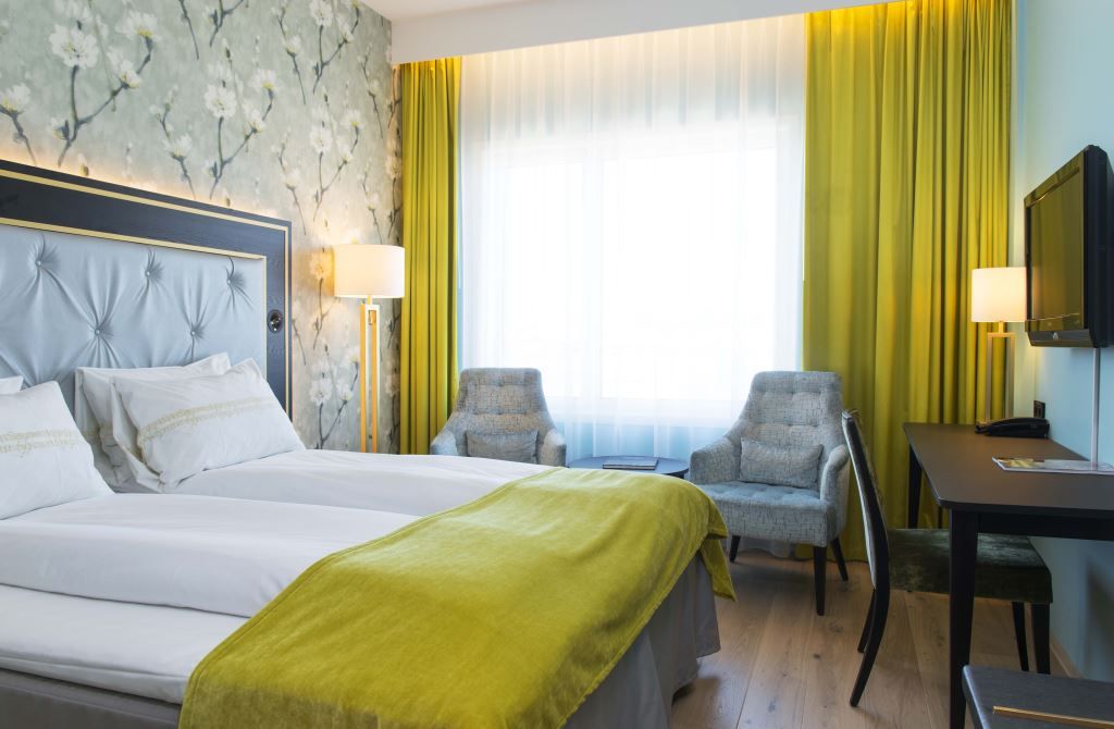 Interiør av hotellrom: Dobbeltseng, blå lenestol, TV-skjerm på veggen, blomstret tapet.