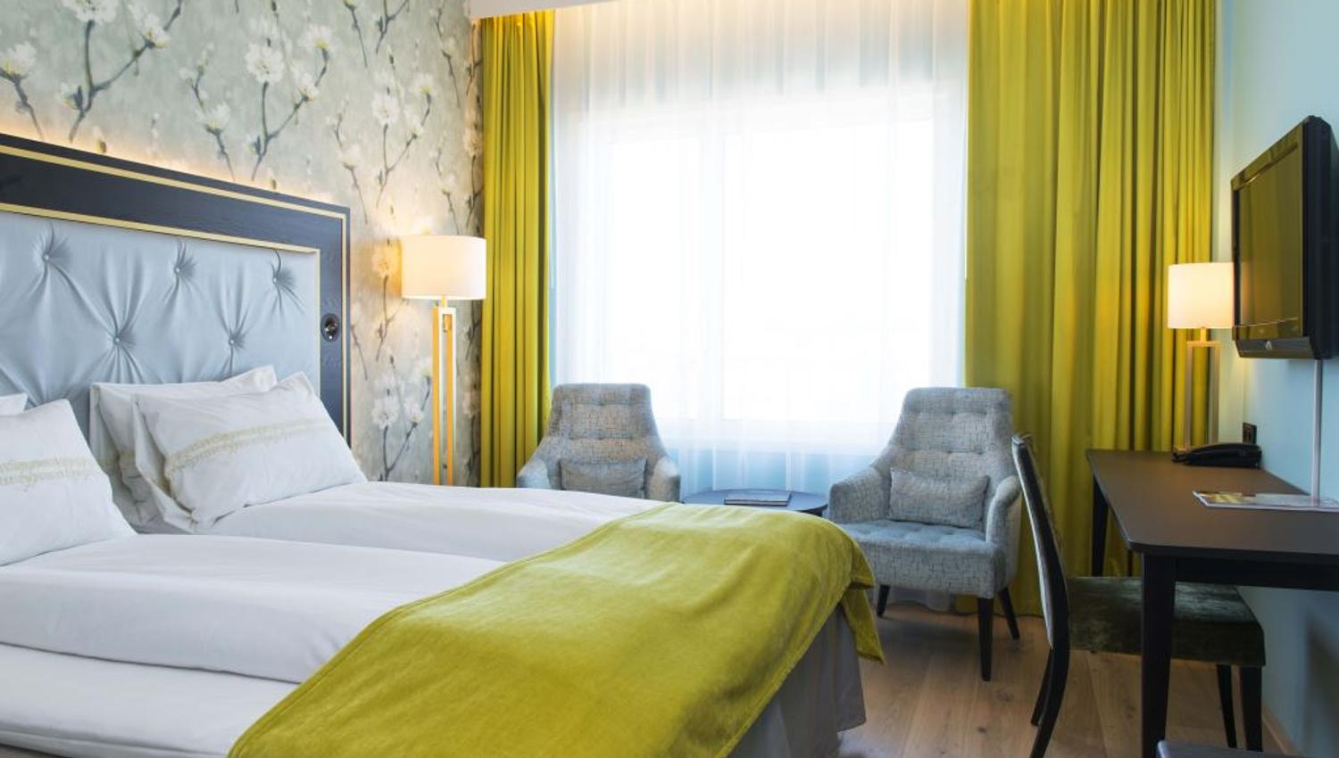 Interiør av hotellrom: Dobbeltseng, blå lenestol, TV-skjerm på veggen, blomstret tapet.
