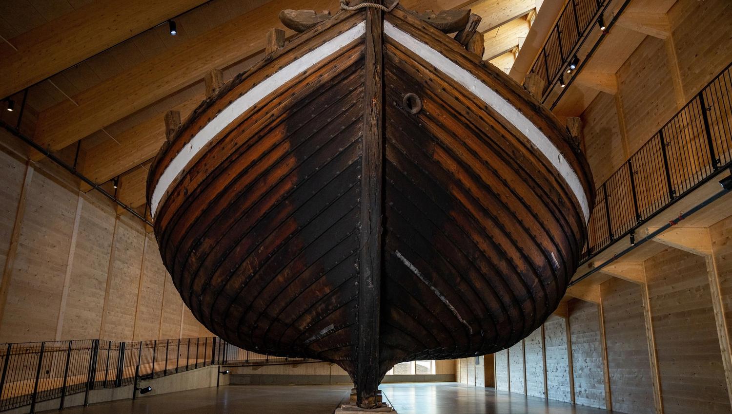 Large wooden boat hull suspended inside a spacious timber-framed hall with railings, spotlights, and polished floor..