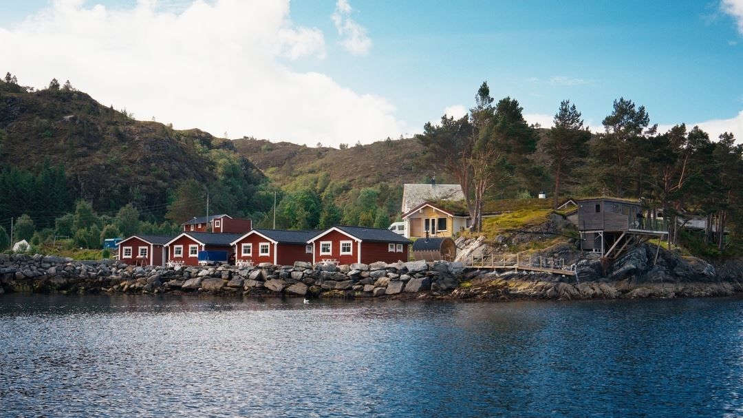 Red cabins and houses line a rocky shoreline beside calm water, with pine trees and hills under a blue sky..