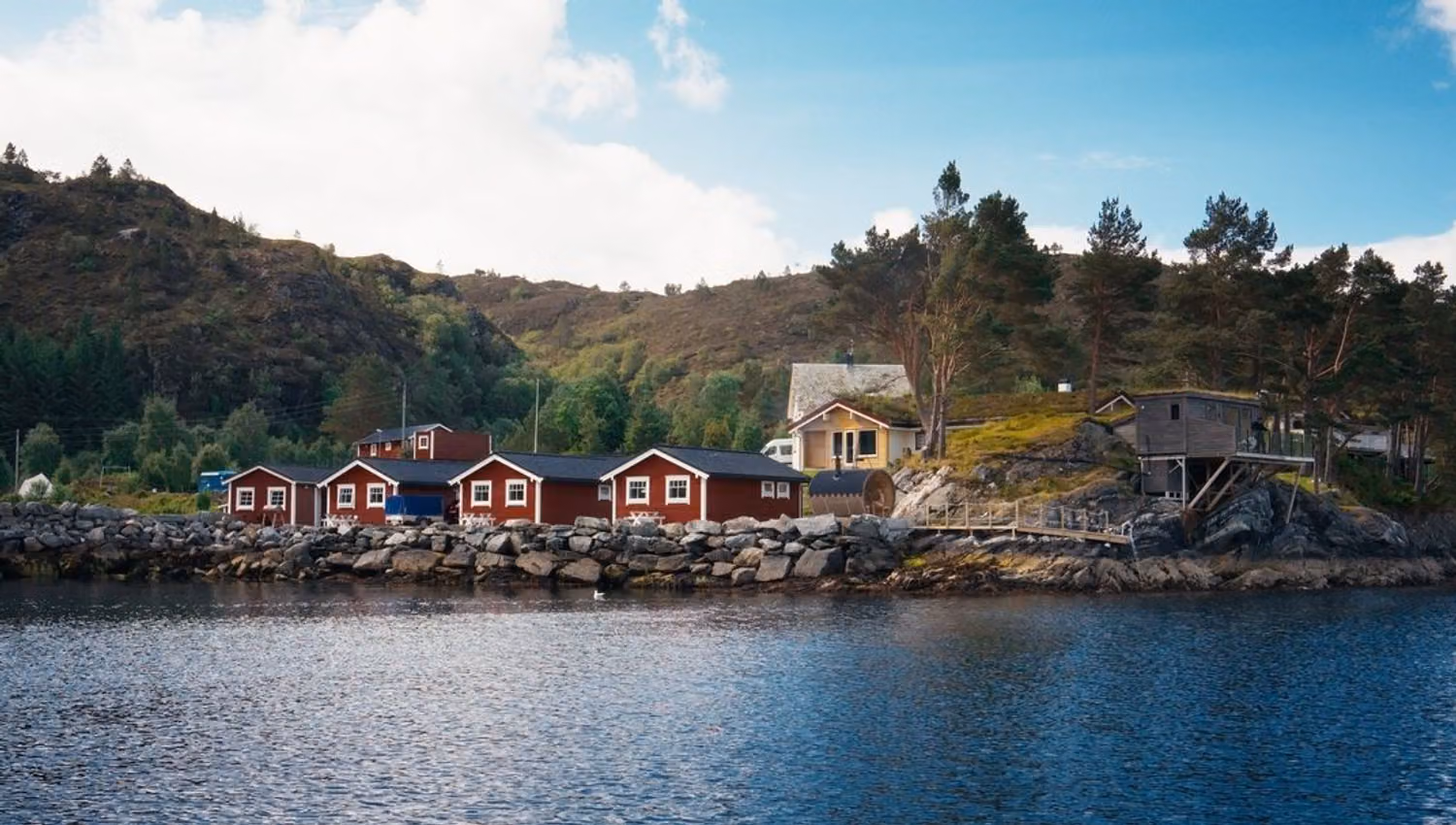 Red cabins and houses line a rocky shoreline beside calm water, with pine trees and hills under a blue sky..