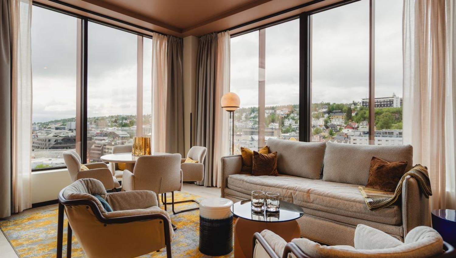 Stue med store vinduer, beige sofa og utsikt mot by og grøntområde