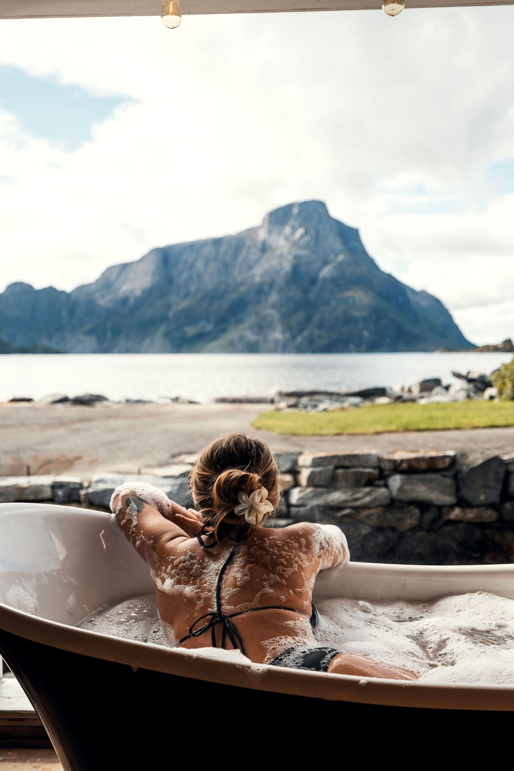 Person reclining in a bubble-filled bathtub on a patio, facing a calm lake and large mountain under clouds at the Seahouse at Hornelen Panorama in Bremanger...