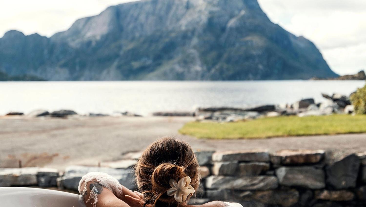 Person reclining in a bubble-filled bathtub on a patio, facing a calm lake and large mountain under clouds at the Seahouse at Hornelen Panorama in Bremanger...