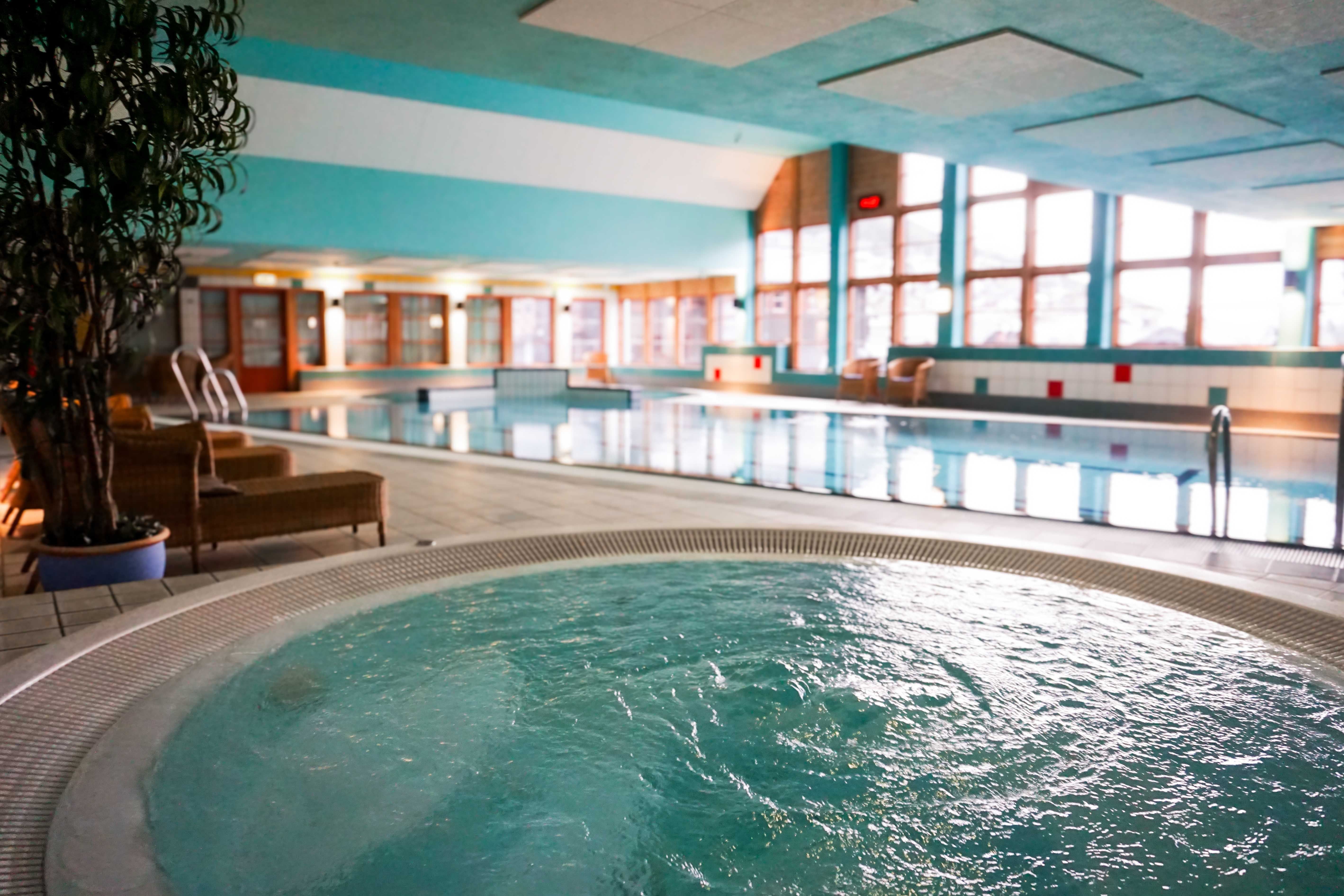 Indoor swimming pool area with a bubbling hot tub in the foreground, lounge chairs, and large windows along the wall..