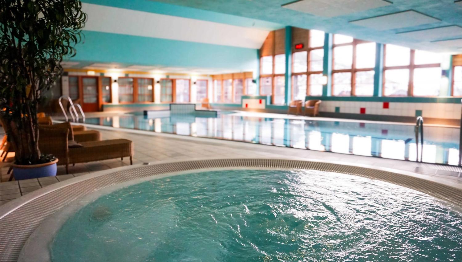 Indoor swimming pool area with a bubbling hot tub in the foreground, lounge chairs, and large windows along the wall..