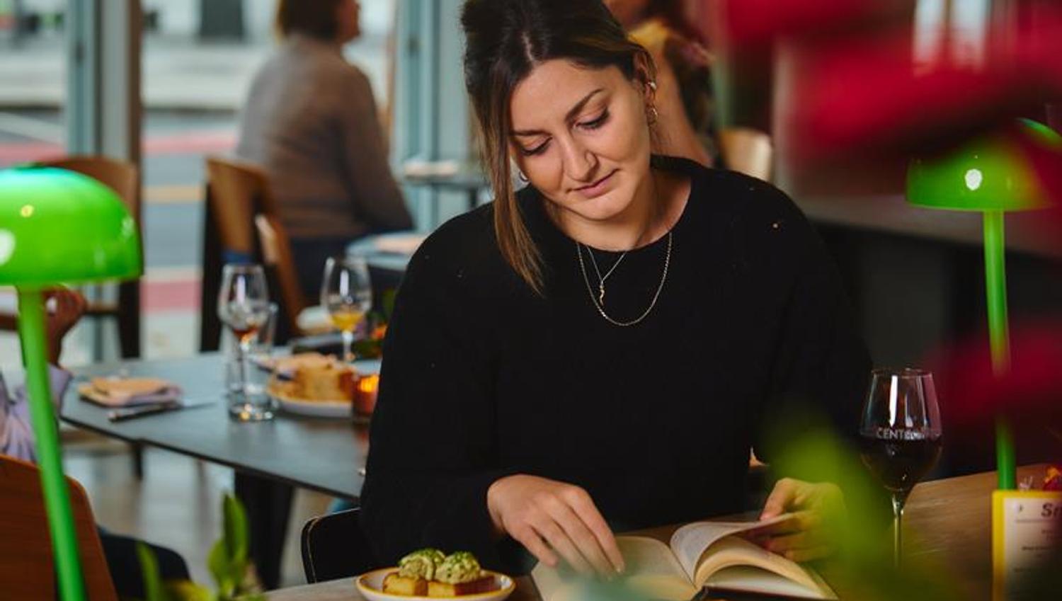 Lady sitting and reading at a café table with food and a glass of wine, flowers, and two green lamps on the table