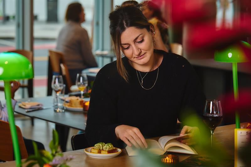 Lady sitting and reading at a café table with food and a glass of wine, flowers, and two green lamps on the table