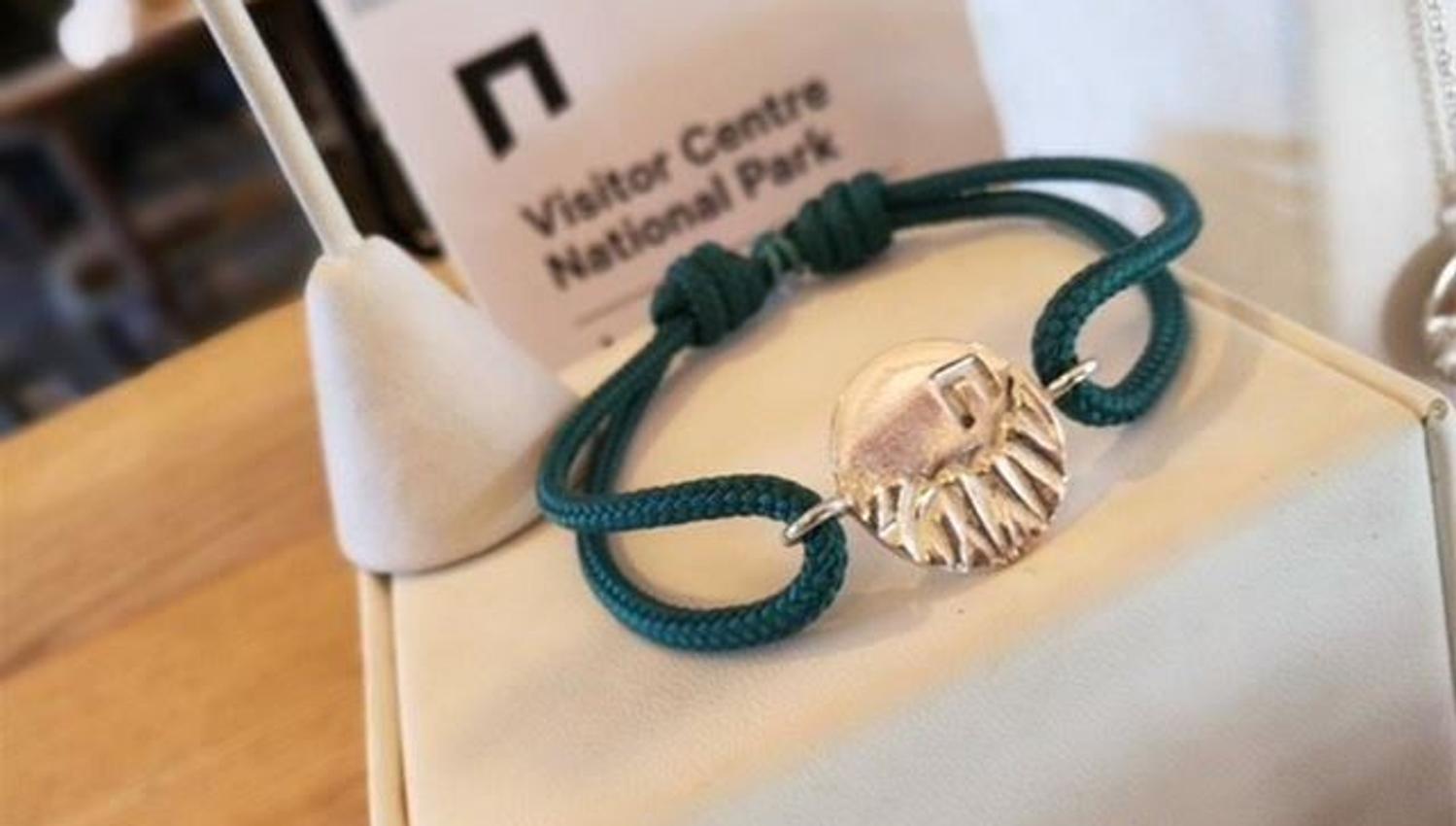 Nærbilete av grønt tauarmband med sølvfarga anheng, lagt på ei lys pute med skilt i bakgrunnen..