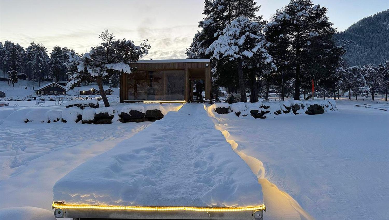 Badstu ved innsjø med opplyst badebrygge om vinteren i dyp snø