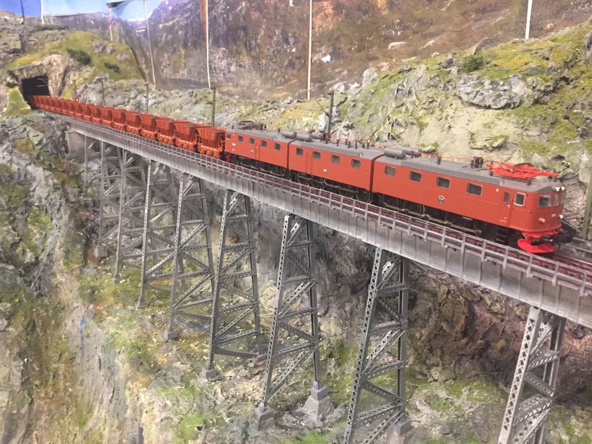 A red electric locomotive hauling freight cars crosses a tall steel trestle bridge over a rocky mountain gorge.