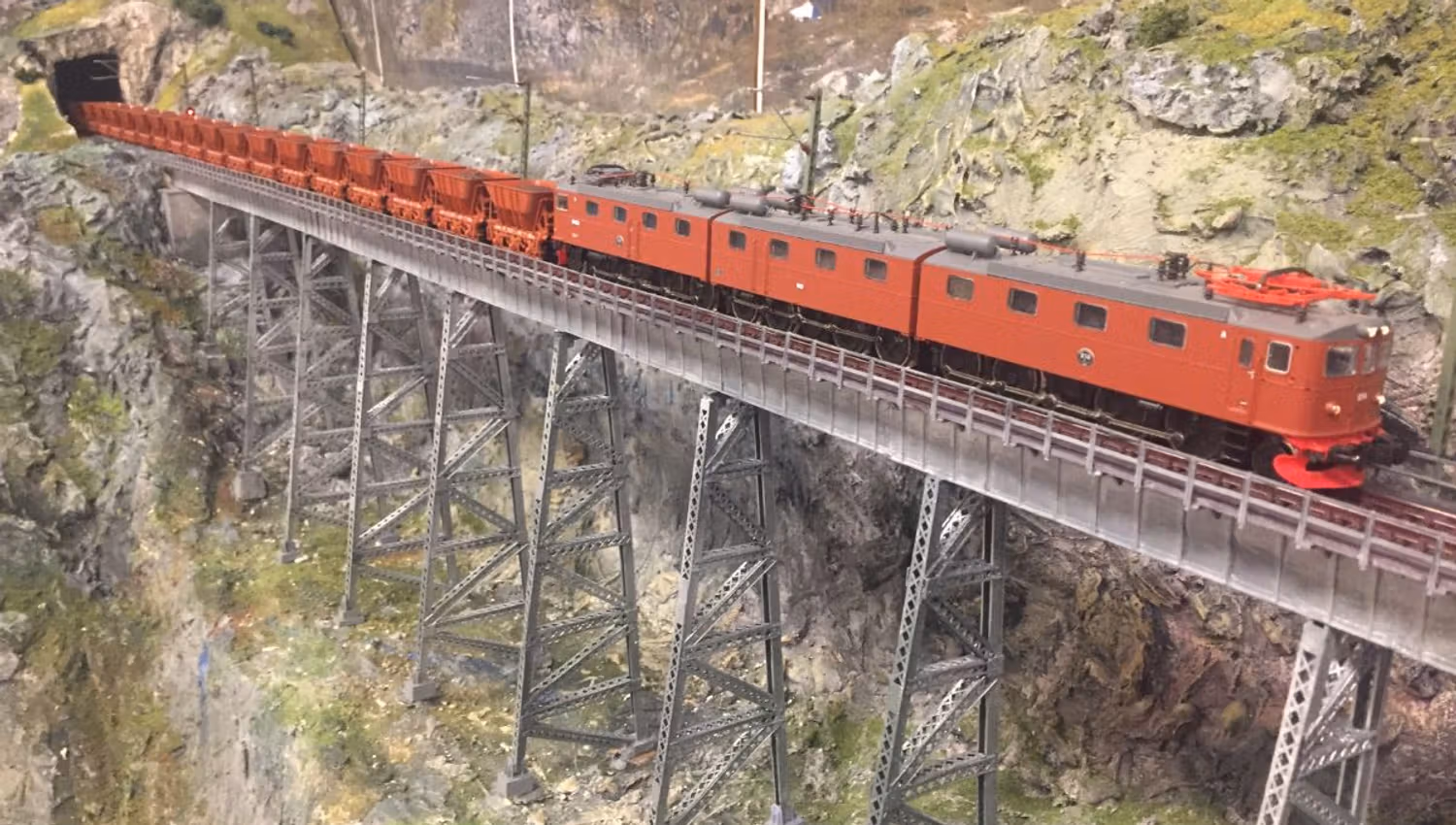 A red electric locomotive hauling freight cars crosses a tall steel trestle bridge over a rocky mountain gorge.
