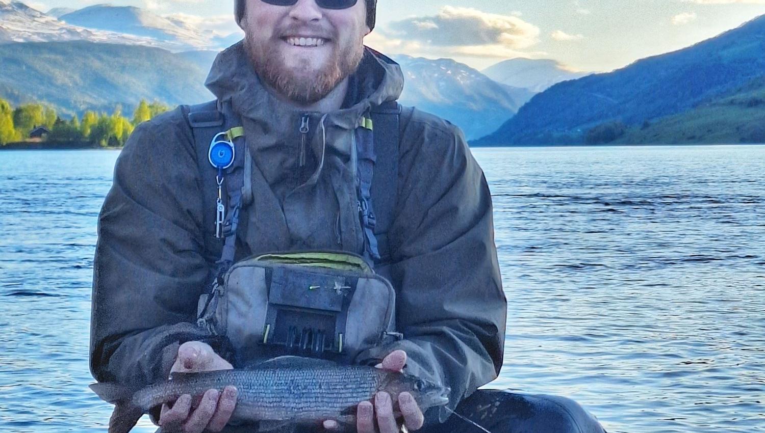 Person holder en fisk ved innsjø, med fjell og vann i bakgrunnen.