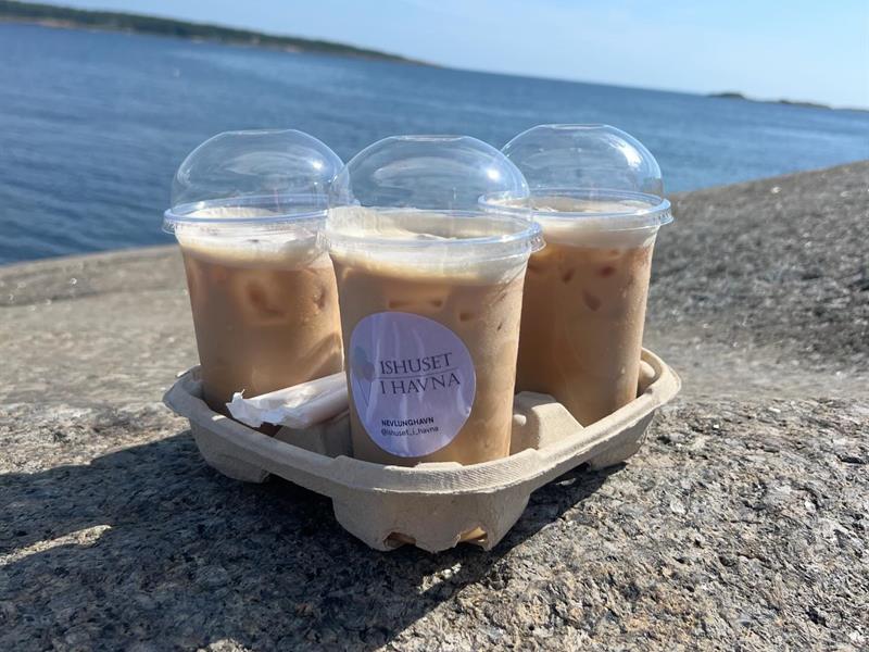 Tre kalde kaffedrikker i plastbegre på et svaberg ved sjøen, med informasjon om dagens åpningstider.