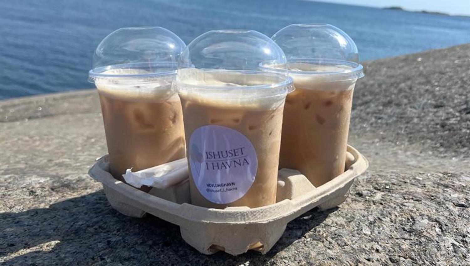Tre kalde kaffedrikker i plastbegre på et svaberg ved sjøen, med informasjon om dagens åpningstider.