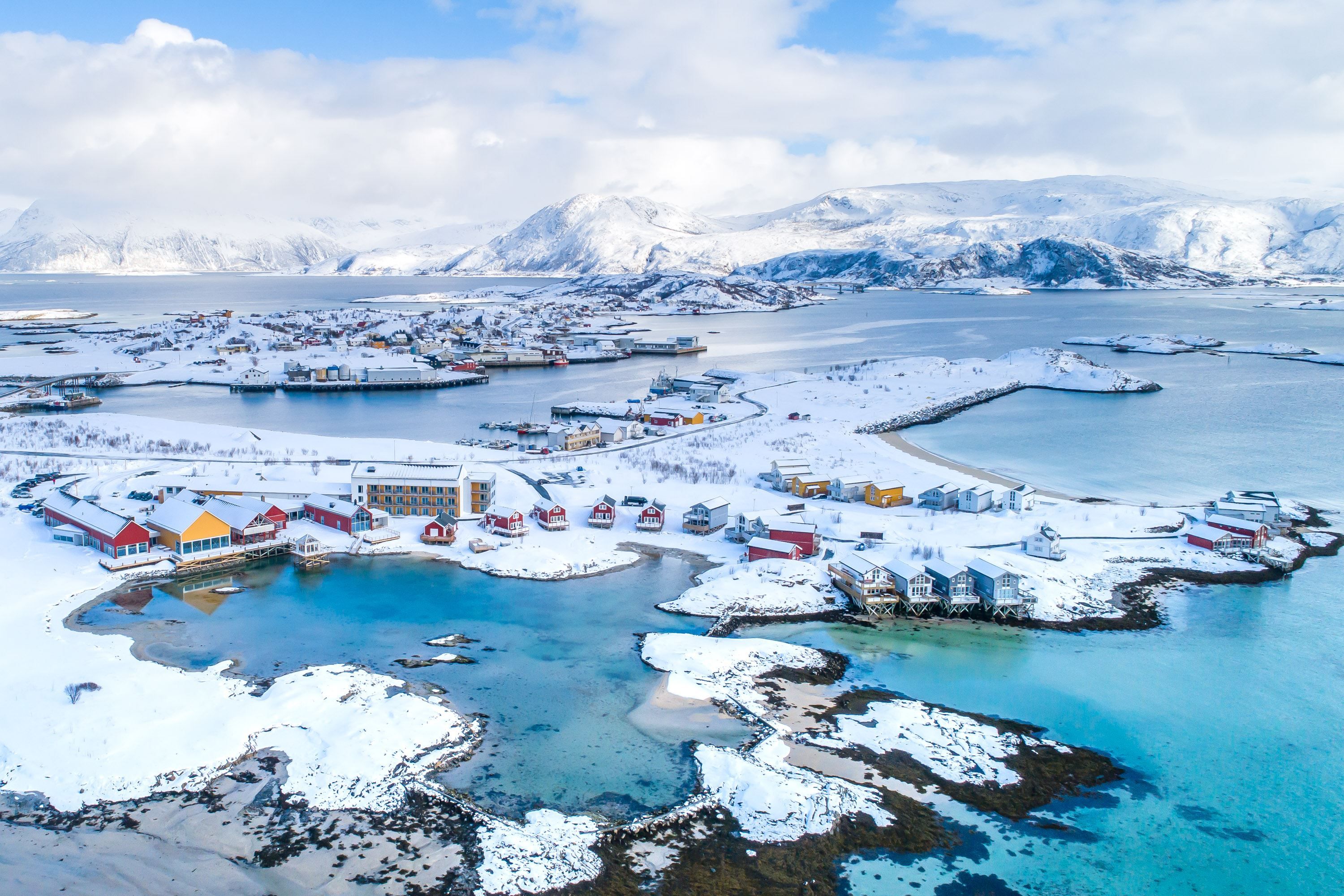Colourful coastal village with snow, mountains, and sea under a partly cloudy sky