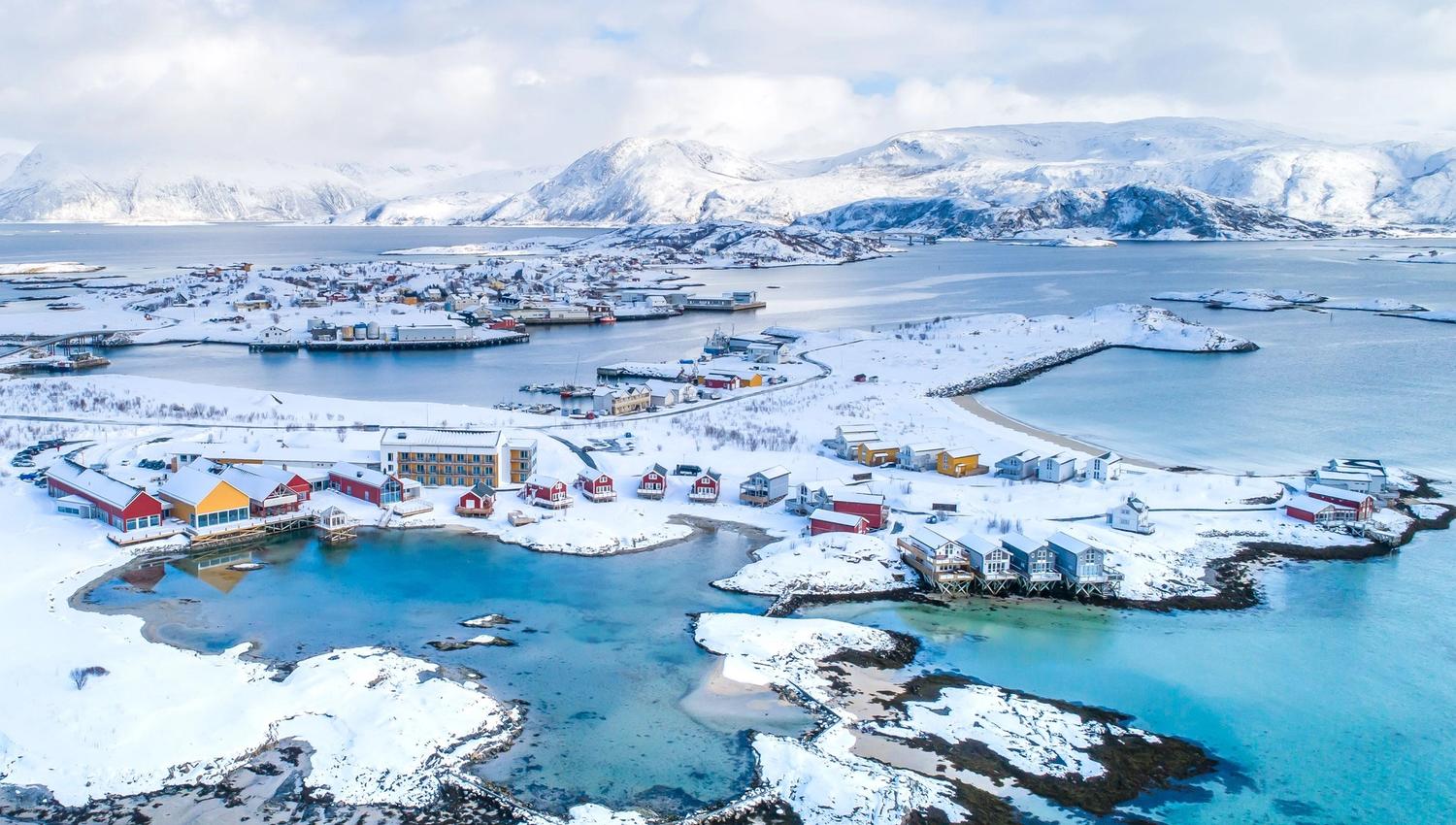 Colourful coastal village with snow, mountains, and sea under a partly cloudy sky