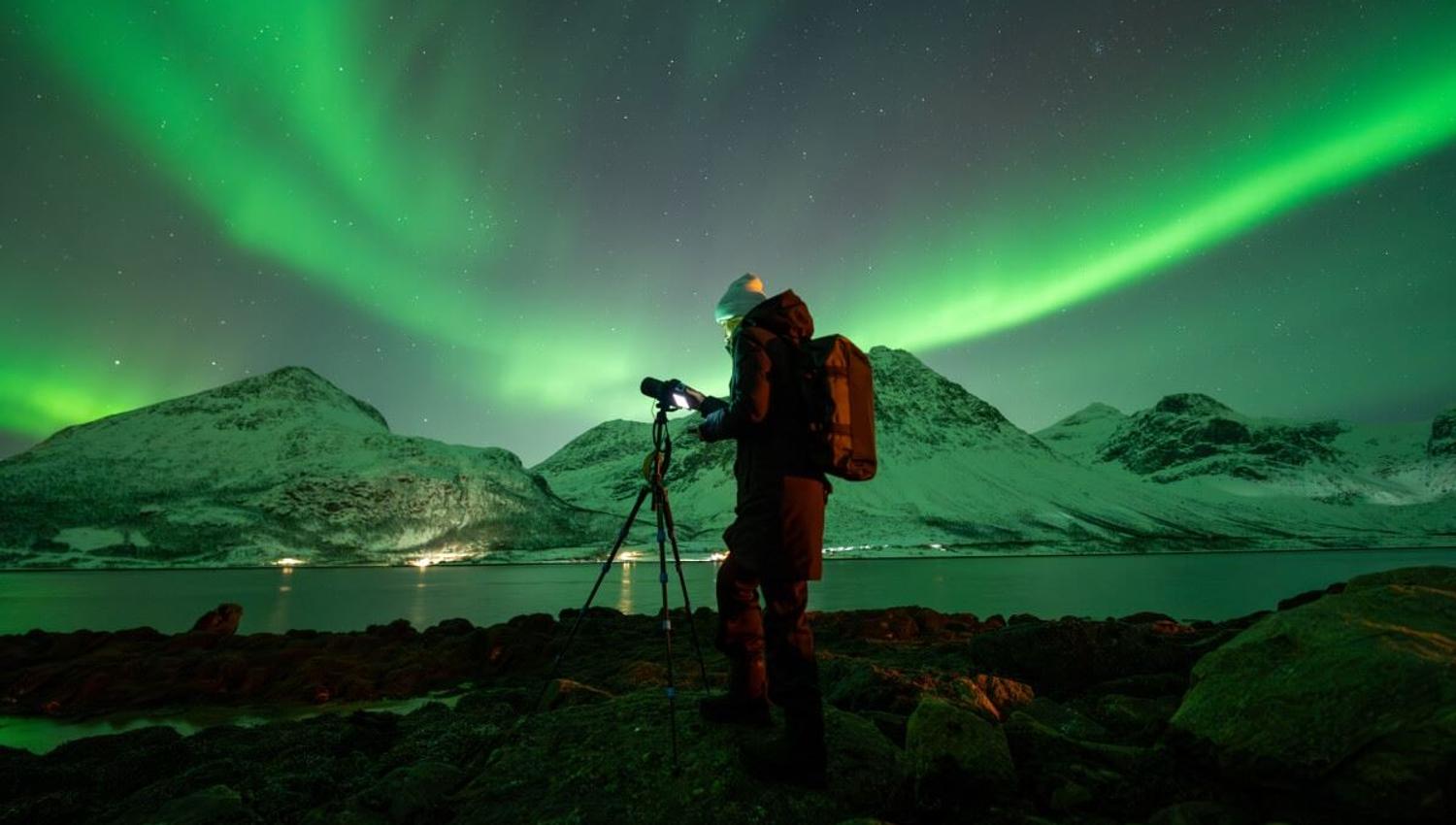 A photographer points the camera up at the Northern Lights, which creates an arch overhead