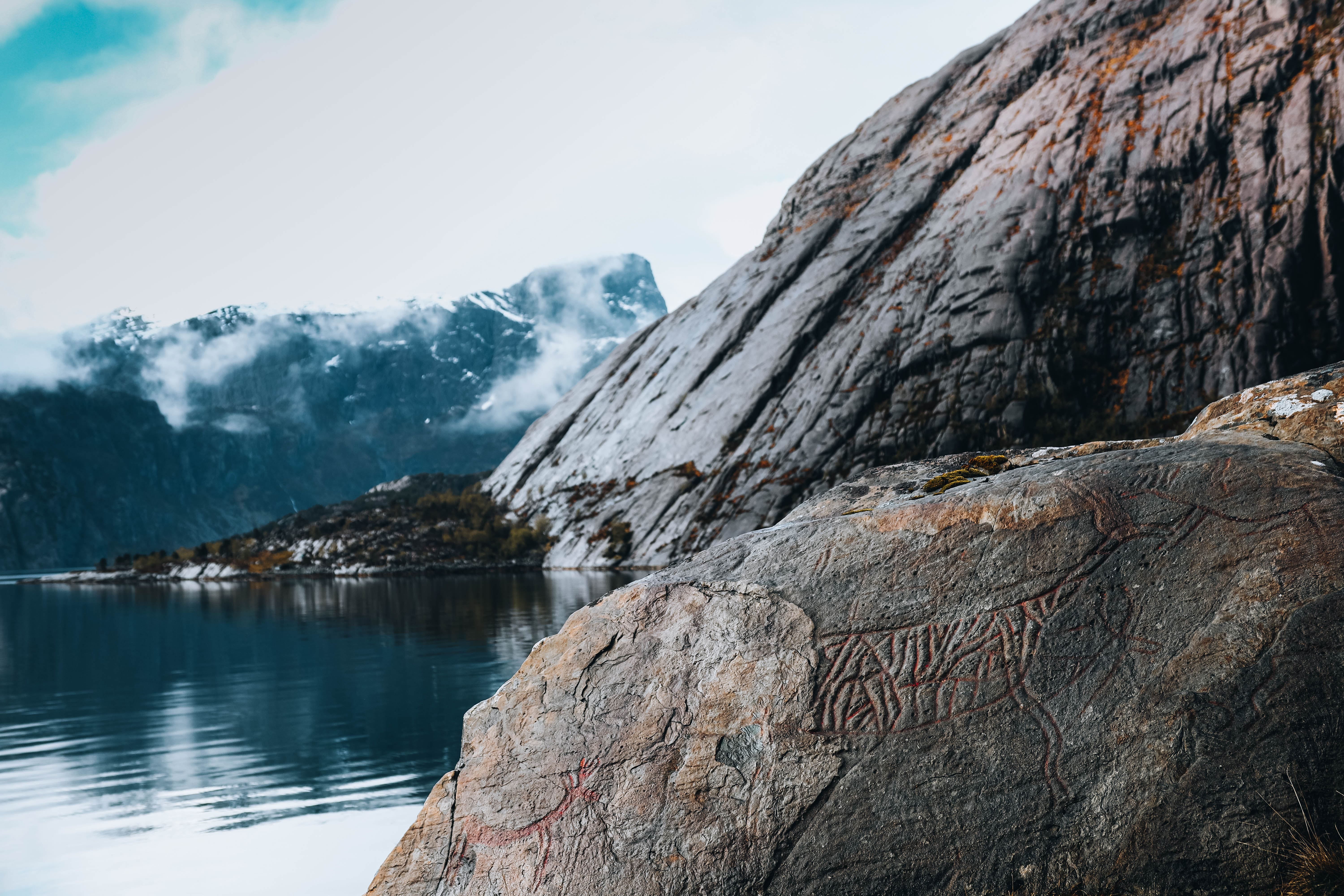 Large weathered rock with faint carvings by a calm lake, with misty mountains and low clouds in the distance.