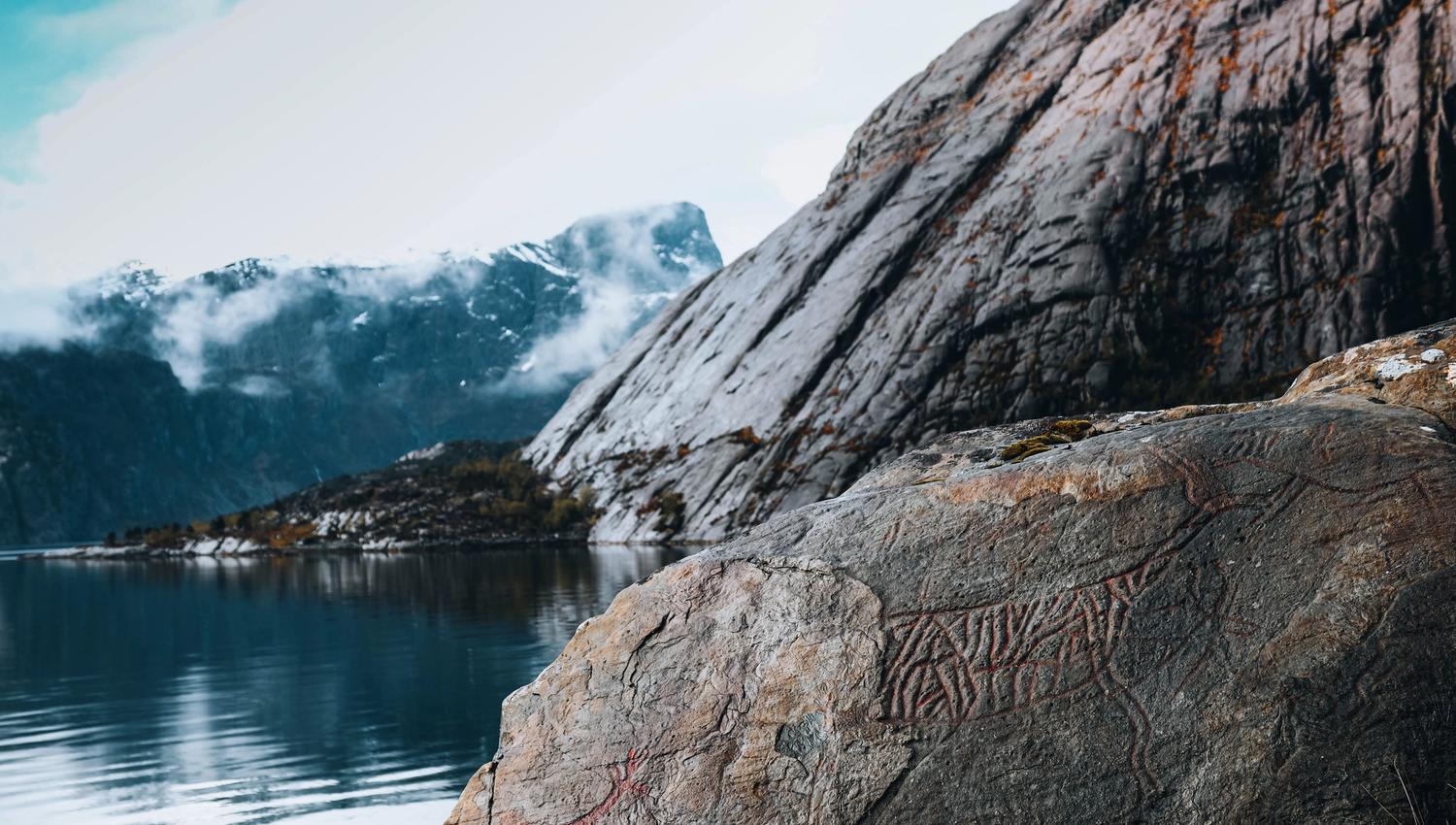 Large weathered rock with faint carvings by a calm lake, with misty mountains and low clouds in the distance.
