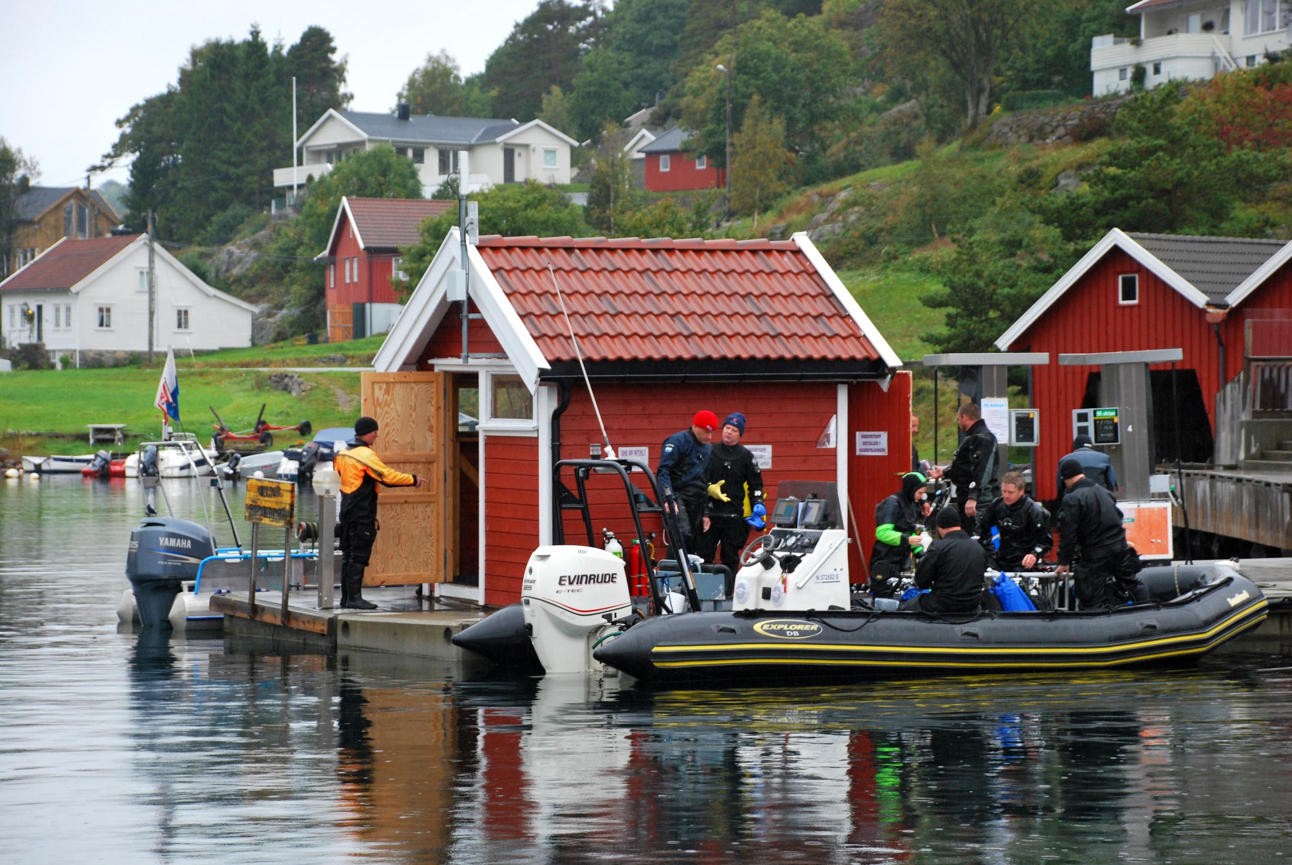 Bild von einer roten Hütte auf einem Pier mit mindestens 8 Tauchern, die sich bereit machen, in ein schwarzes Boot in Tregde zu steigen