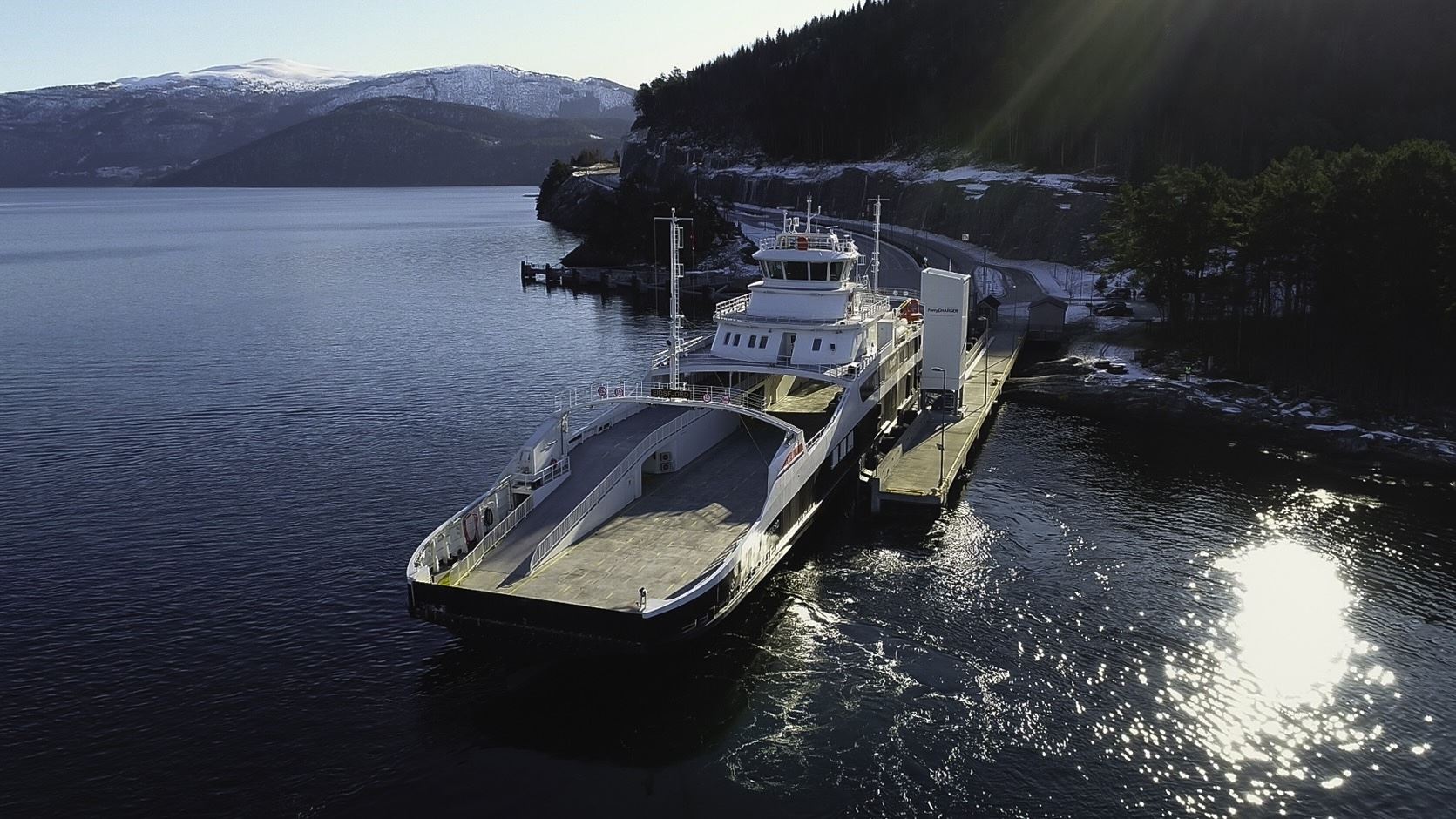 A large car ferry is docked at a pier on a calm fjord, with forested hills and snowy mountains behind..