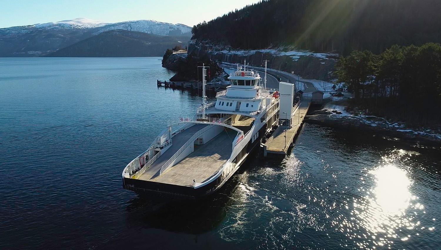 A large car ferry is docked at a pier on a calm fjord, with forested hills and snowy mountains behind..