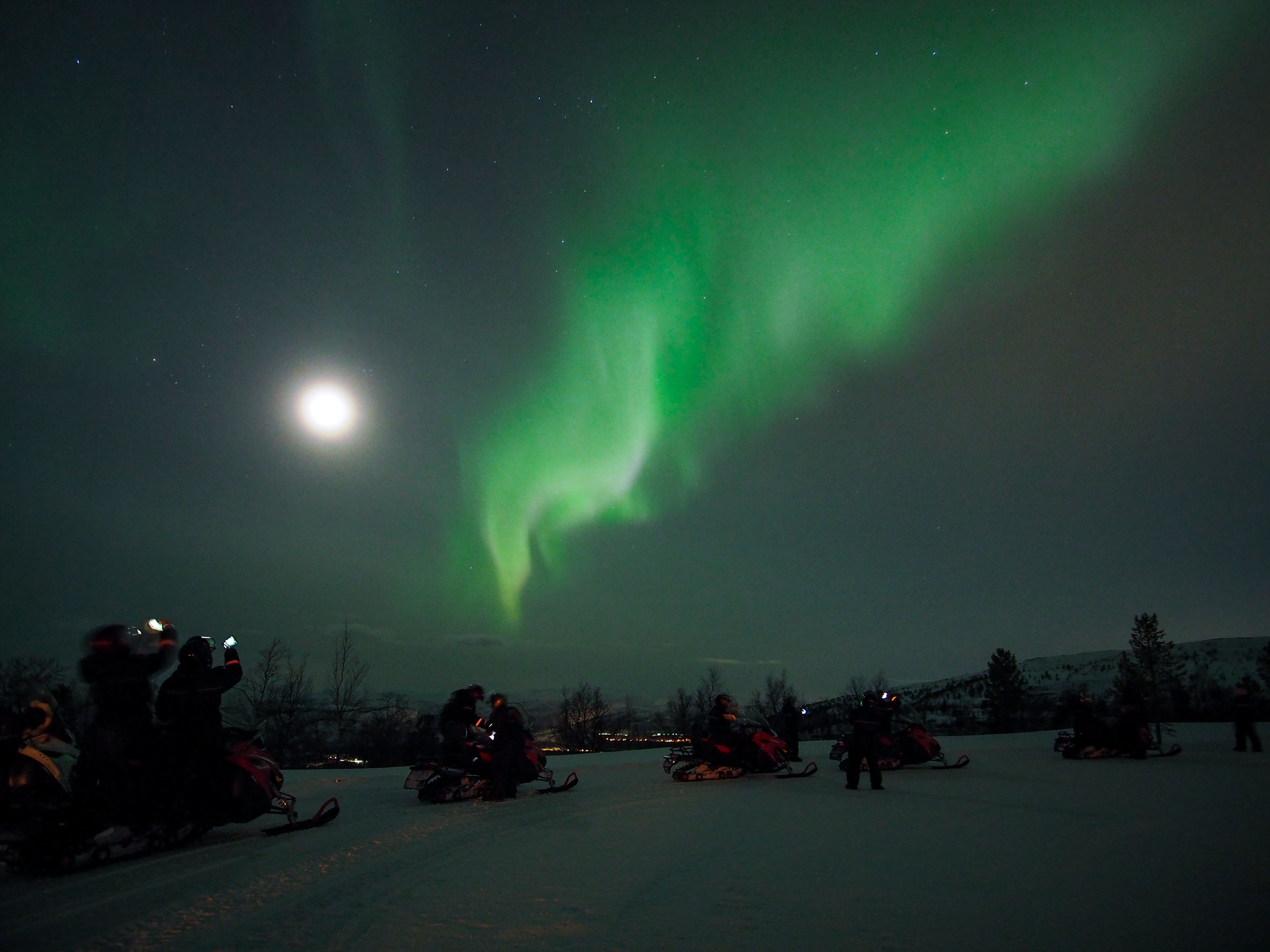 Experience the breathtaking Northern Lights illuminating the night sky, as enthusiastic spectators gather on snowmobiles for an unforgettable adventure in a wintry wonderland.