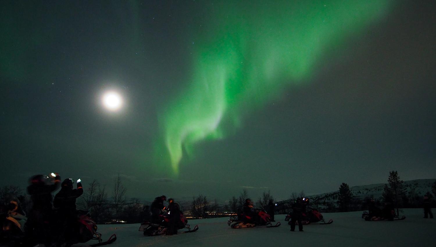 Experience the breathtaking Northern Lights illuminating the night sky, as enthusiastic spectators gather on snowmobiles for an unforgettable adventure in a wintry wonderland.