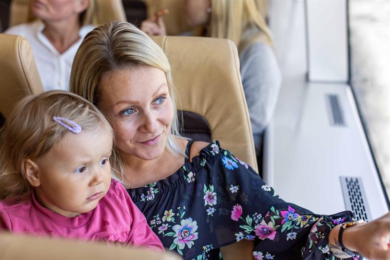 Mor og lite barn sit i komfortable salongstoler innandørs om bord på fjordcruisebåten på Hardangerfjorden.