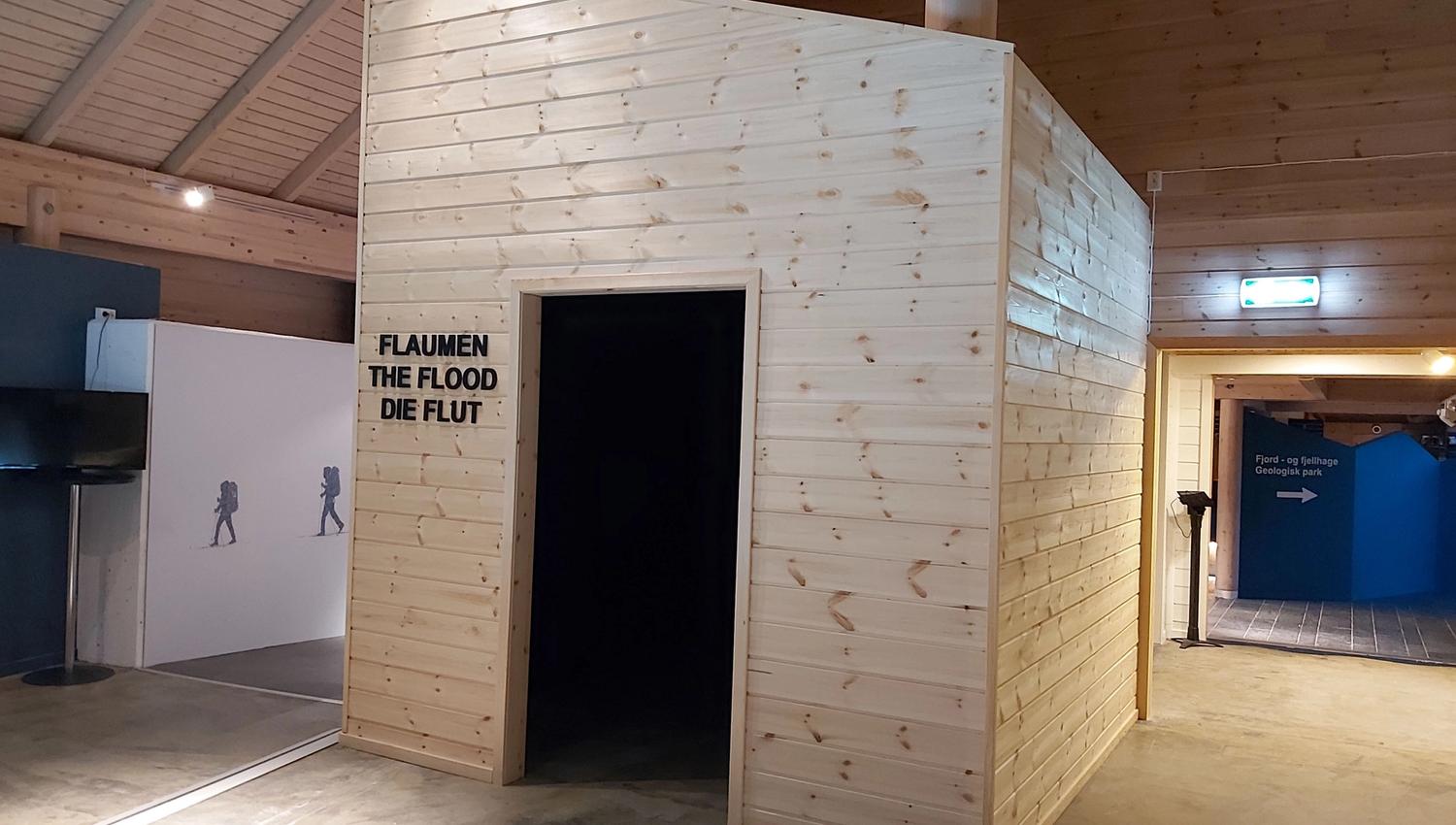 Innandørs utstilling med stor treboks, open dør og veggtekst «FLAUMEN / THE FLOOD / DIE FLUT» i eit trebygg..