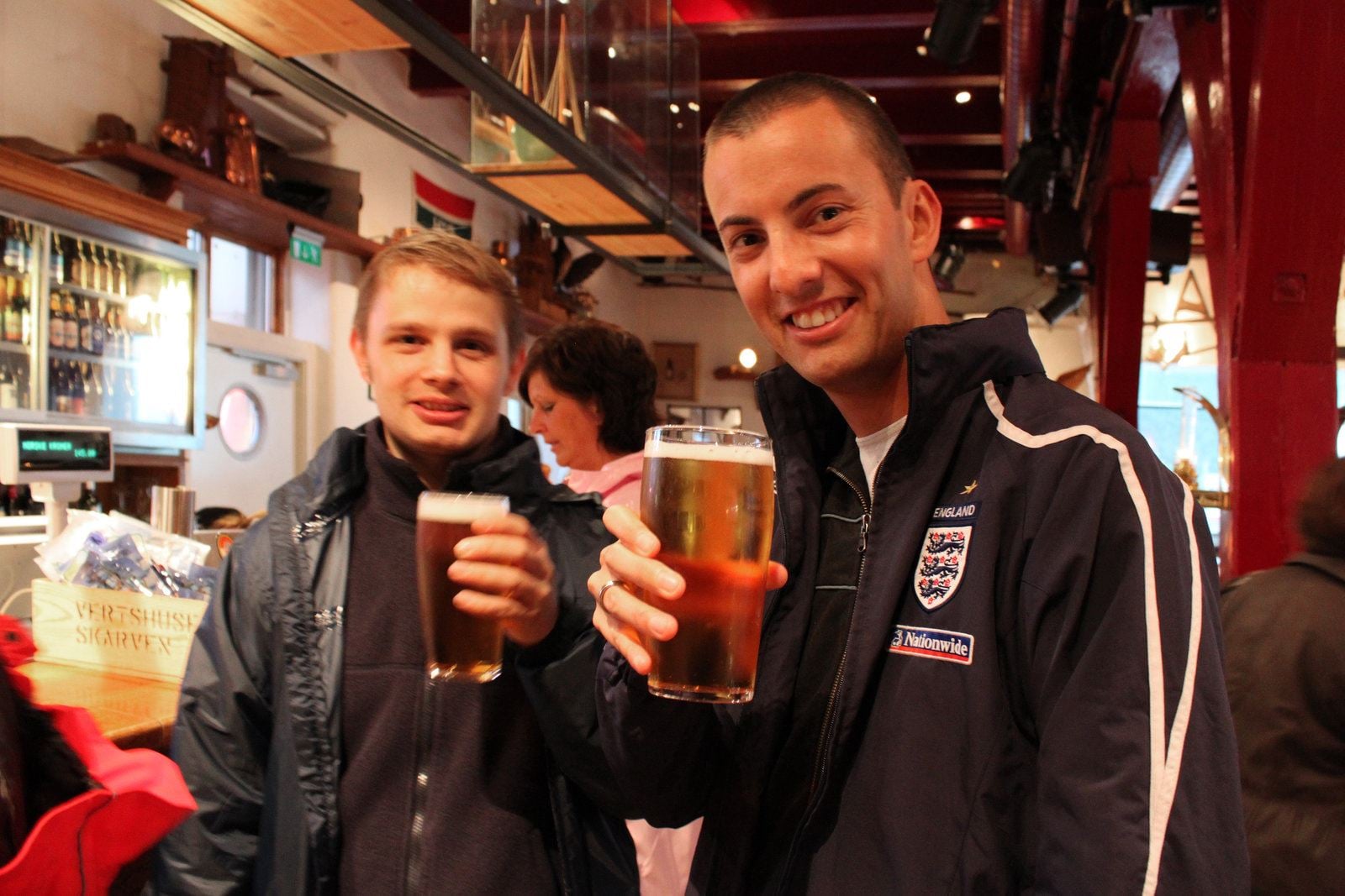 Two people toasting with beer in a bar, one wearing an England jacket.