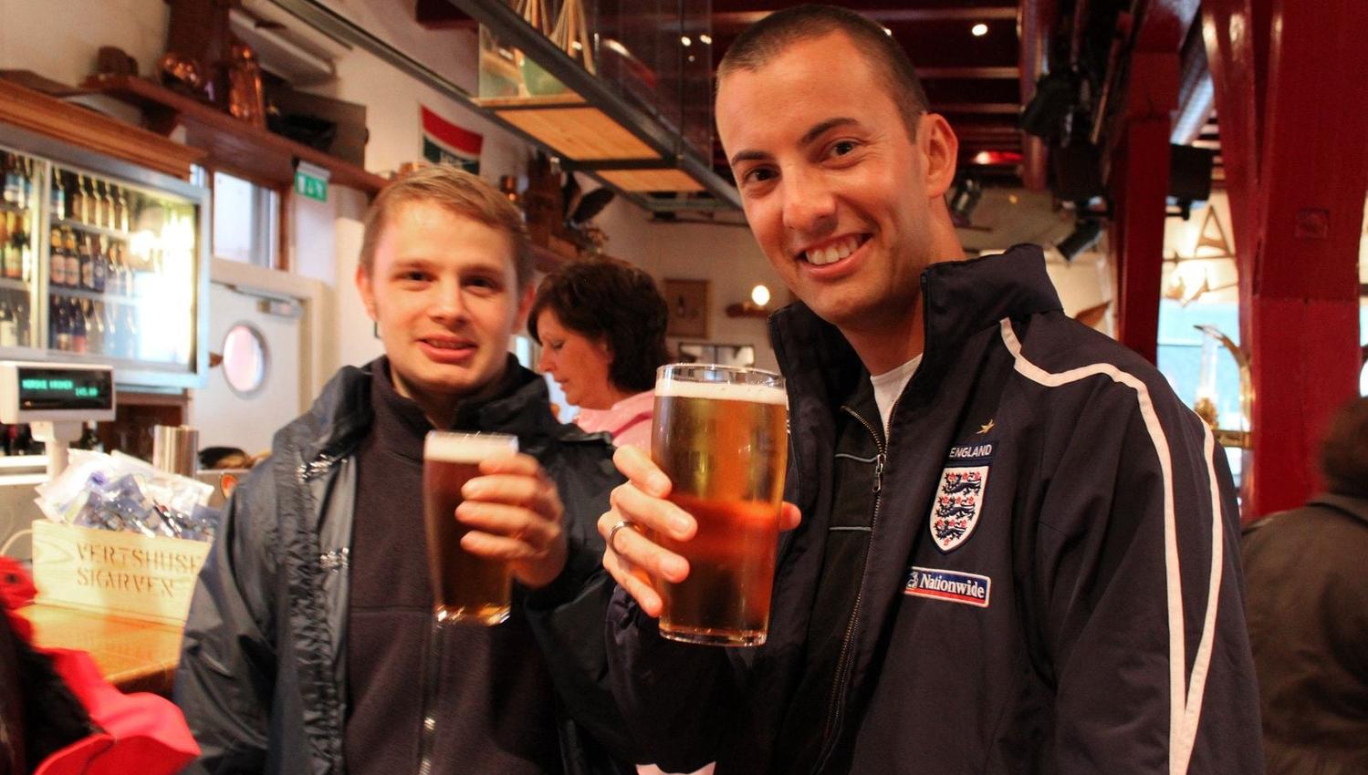 Two people toasting with beer in a bar, one wearing an England jacket.