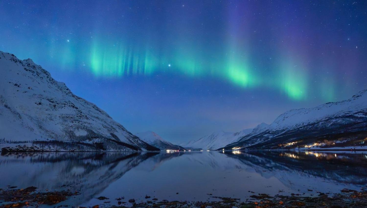 Green Northern Lights illuminate the night sky above an Arctic fjord and snow-covered mountains in Northern Norway. A natural phenomenon guests may be lucky enough to witness during their Northern Lights chas