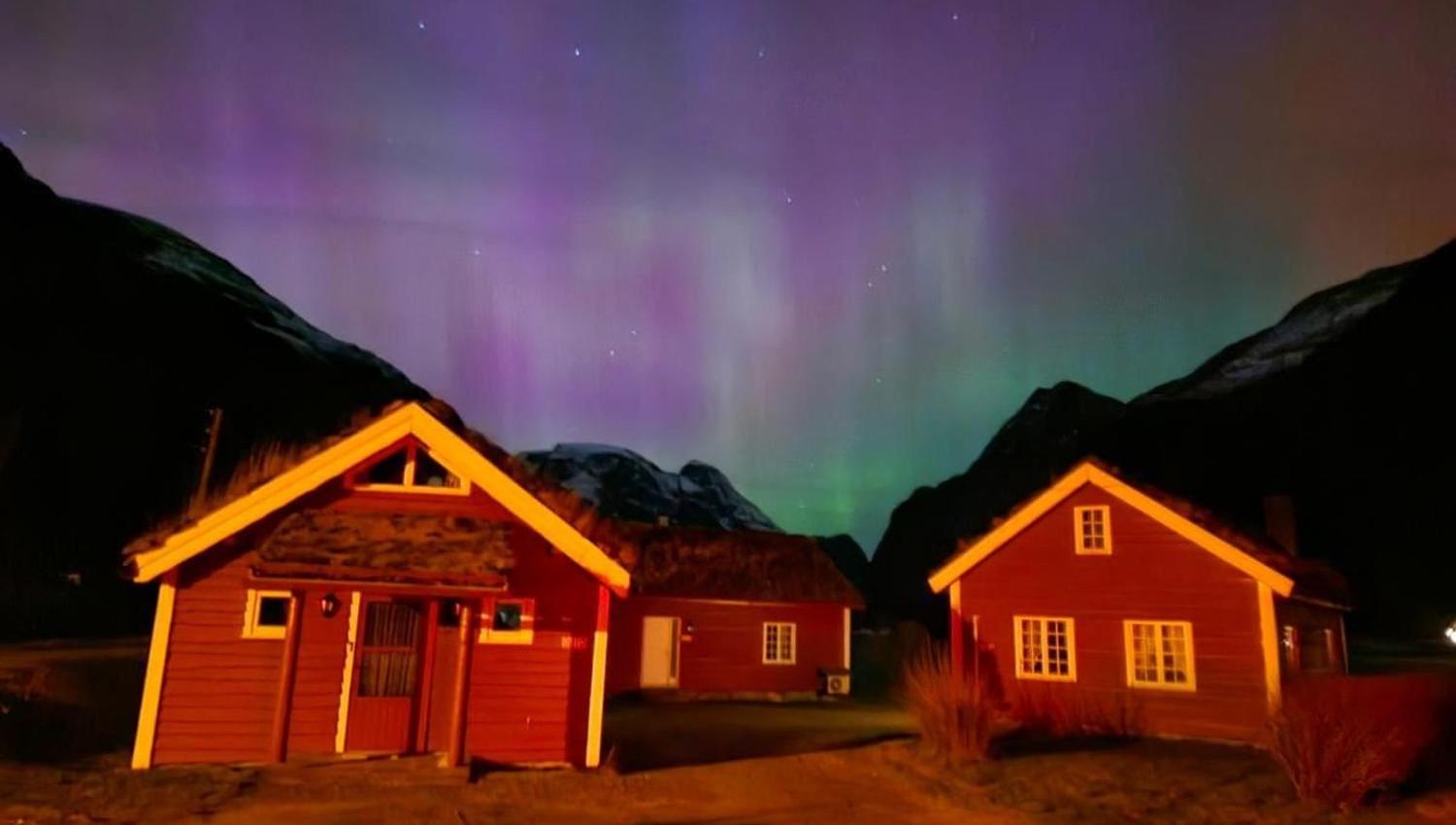 Two red cabins lit in warm light beneath a purple and green aurora sky, framed by dark mountains at night at Aabrekk Gard Trollbu in Oldedalen valley..