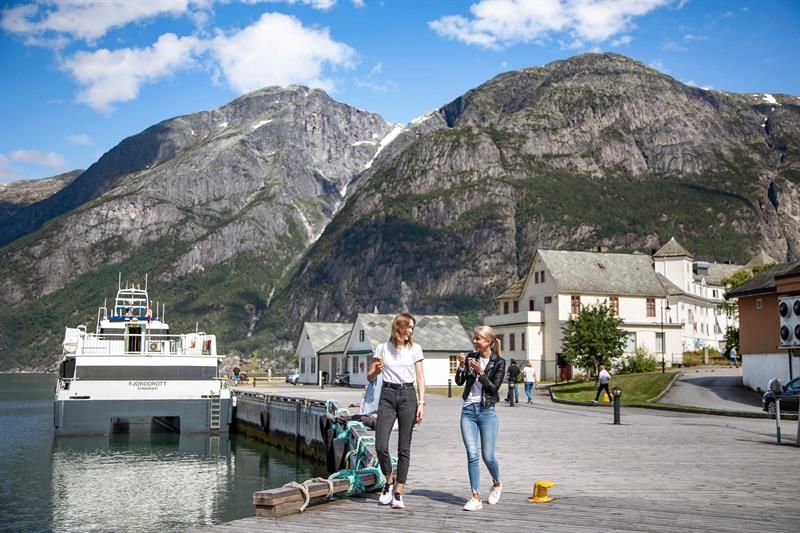 To kvinner går på kaia i Eidfjord med cruisebåten Fjorddrott fortøydd bak og bratte fjell i bakgrunnen.