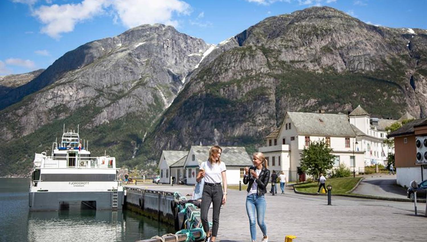 To kvinner går på kaia i Eidfjord med cruisebåten Fjorddrott fortøydd bak og bratte fjell i bakgrunnen.