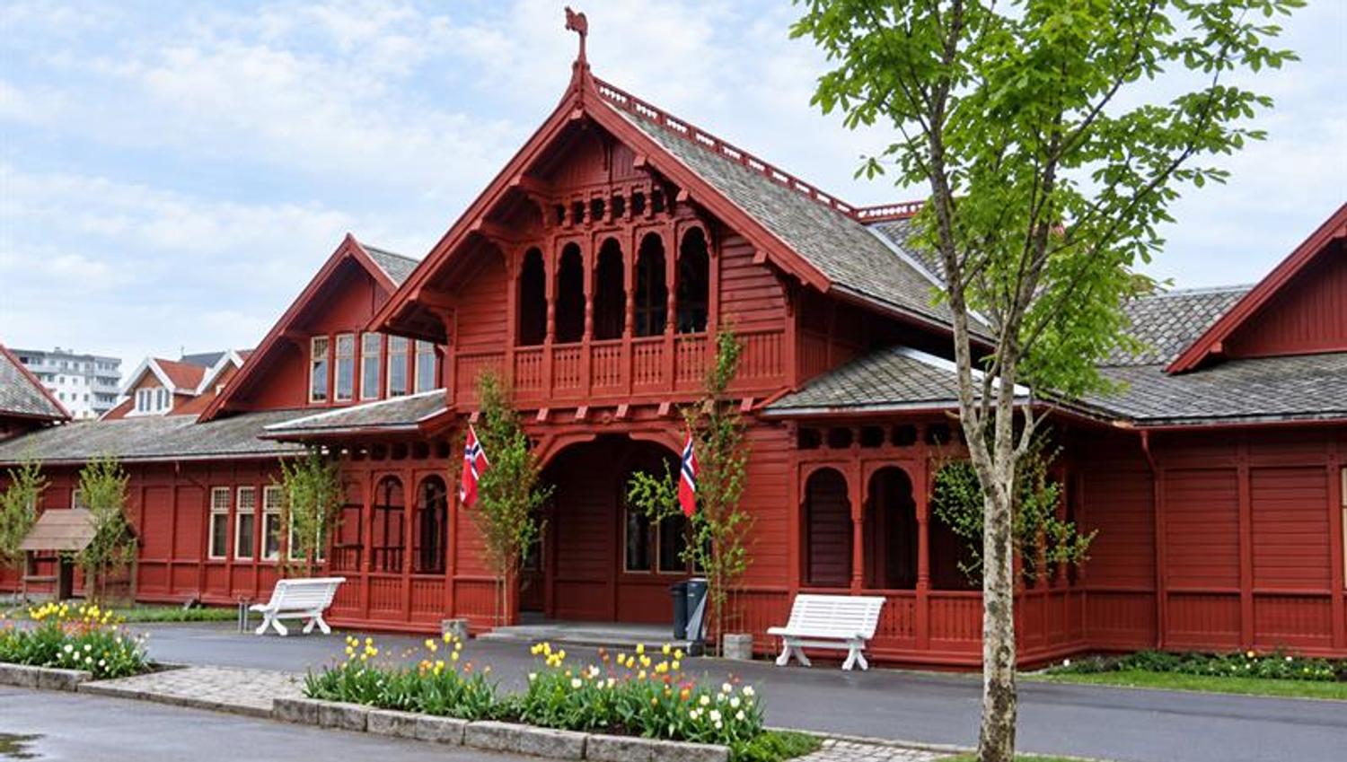 Historic red wooden spa building in Sandefjord with ornate wood carvings, Norwegian flags and flower beds by the entrance.