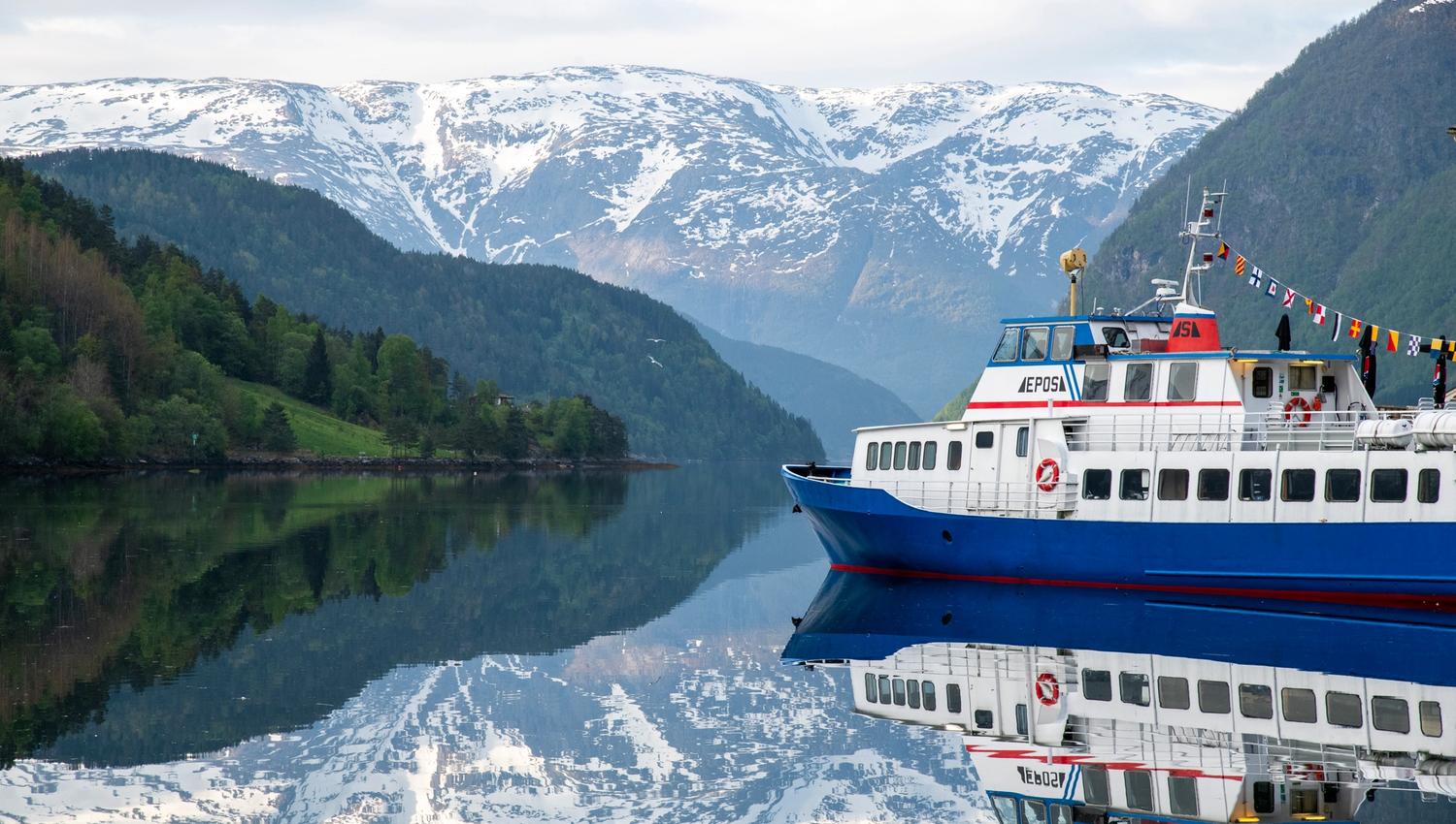 The literature boat Epos resting on the calm Hardangerfjord, surrounded by snow-capped mountains and mirror-like water.